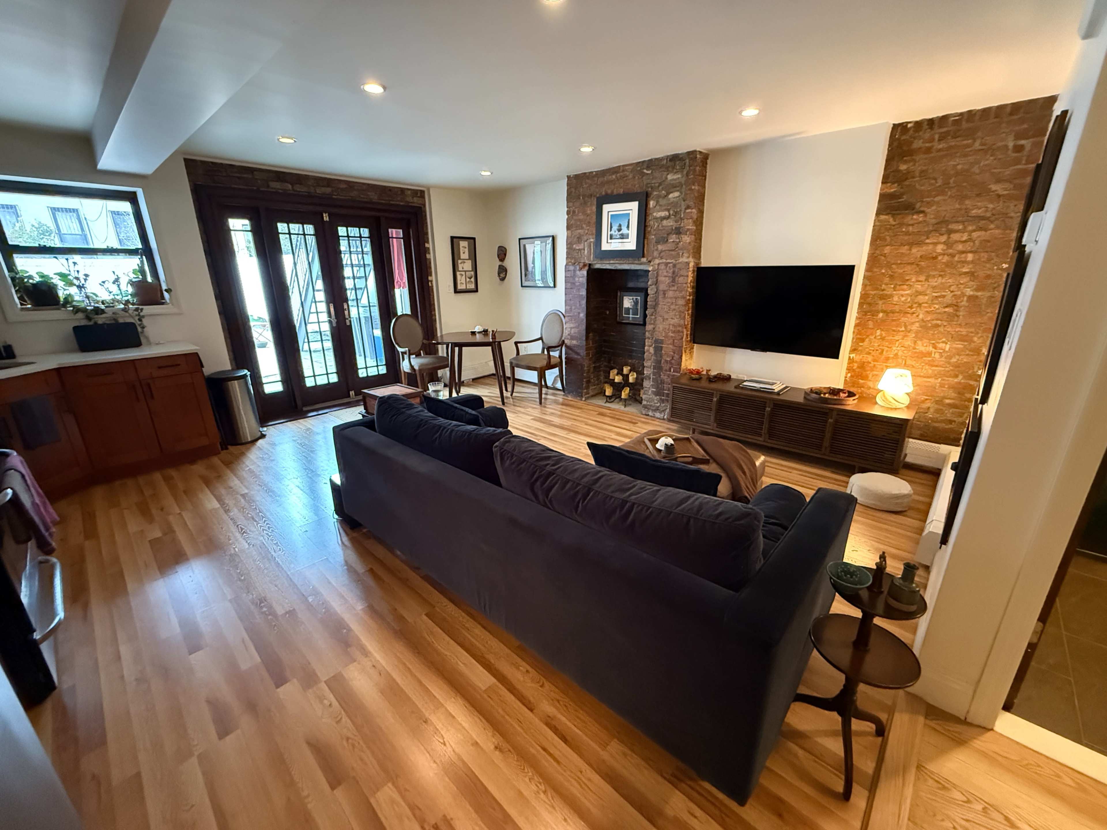 The image shows a modern living room with a dark sofa, a wooden floor, a brick wall, a television, and a dining area with a round table and chairs.