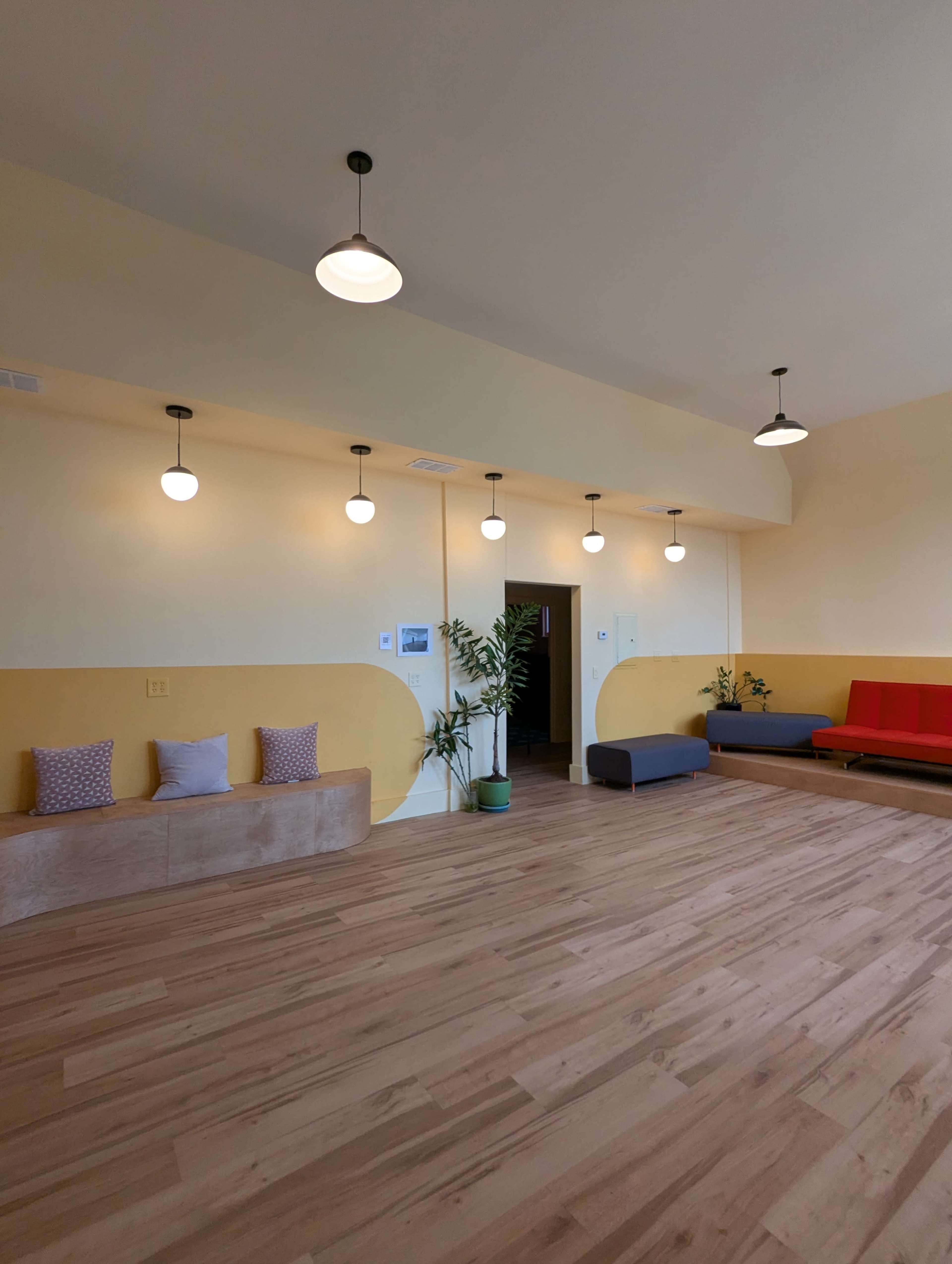 The image shows a spacious room with light wood flooring, yellow and white walls, and several pendant lights, along with seating areas featuring benches and couches.