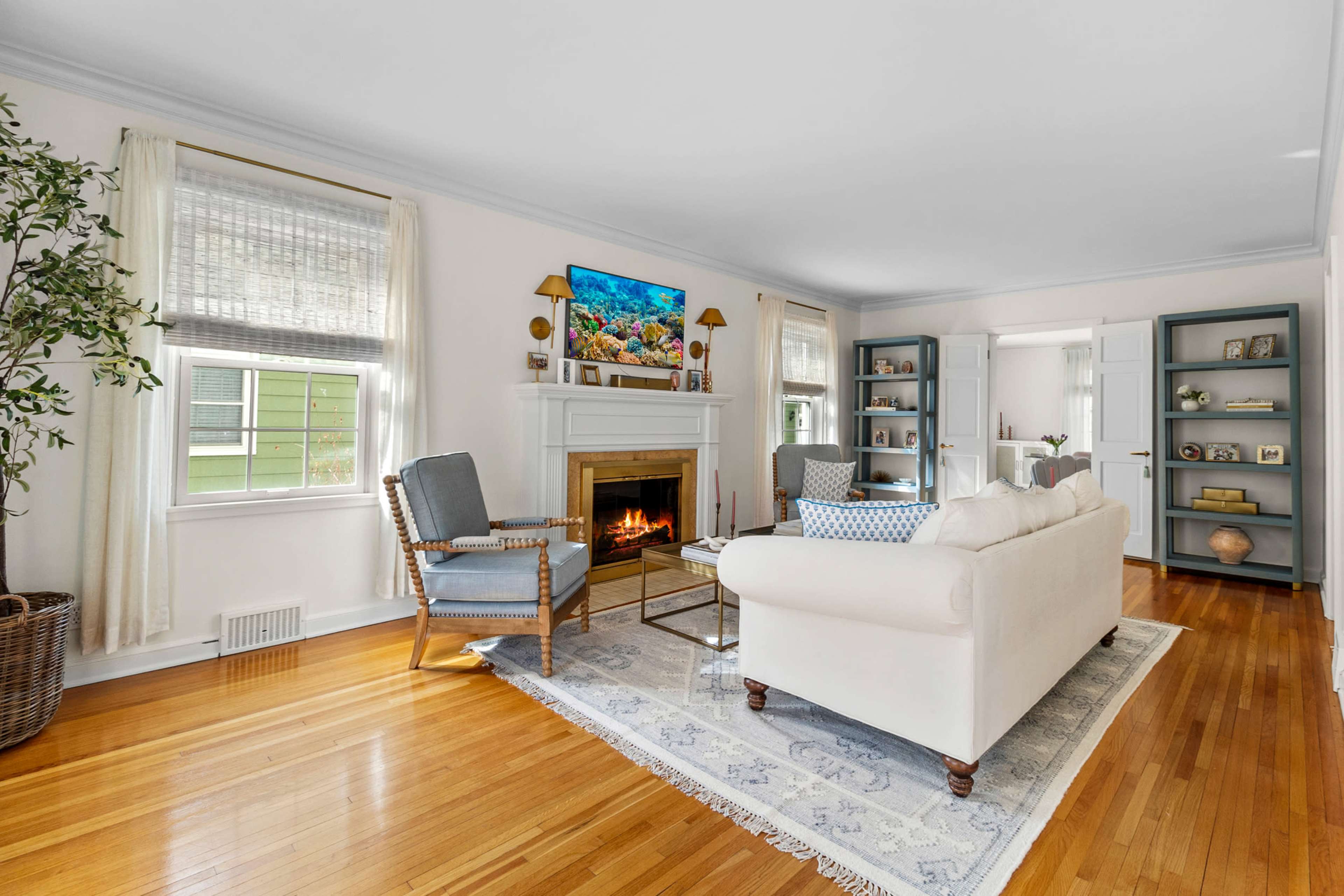 A bright living room features a white sofa facing a fireplace, flanked by two chairs, with shelves and a television on the walls.