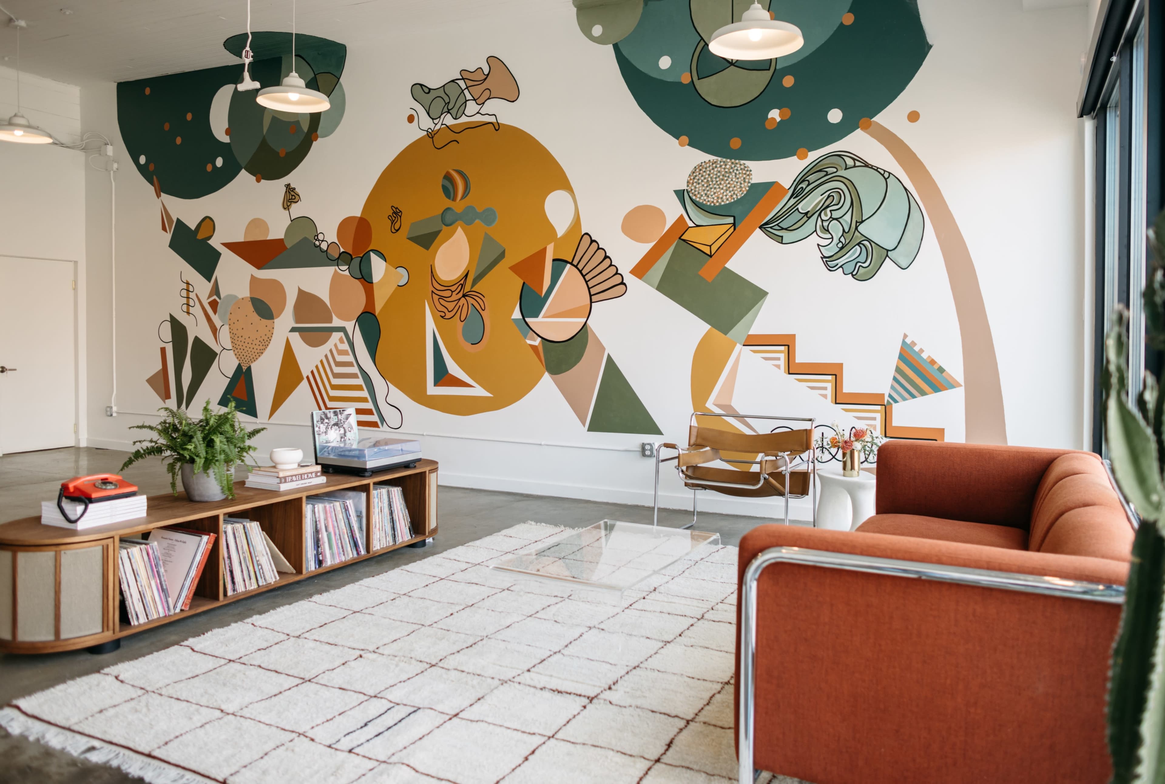 A bright interior space featuring a colorful mural, a modern orange sofa, a wooden coffee table with books, and a patterned rug.