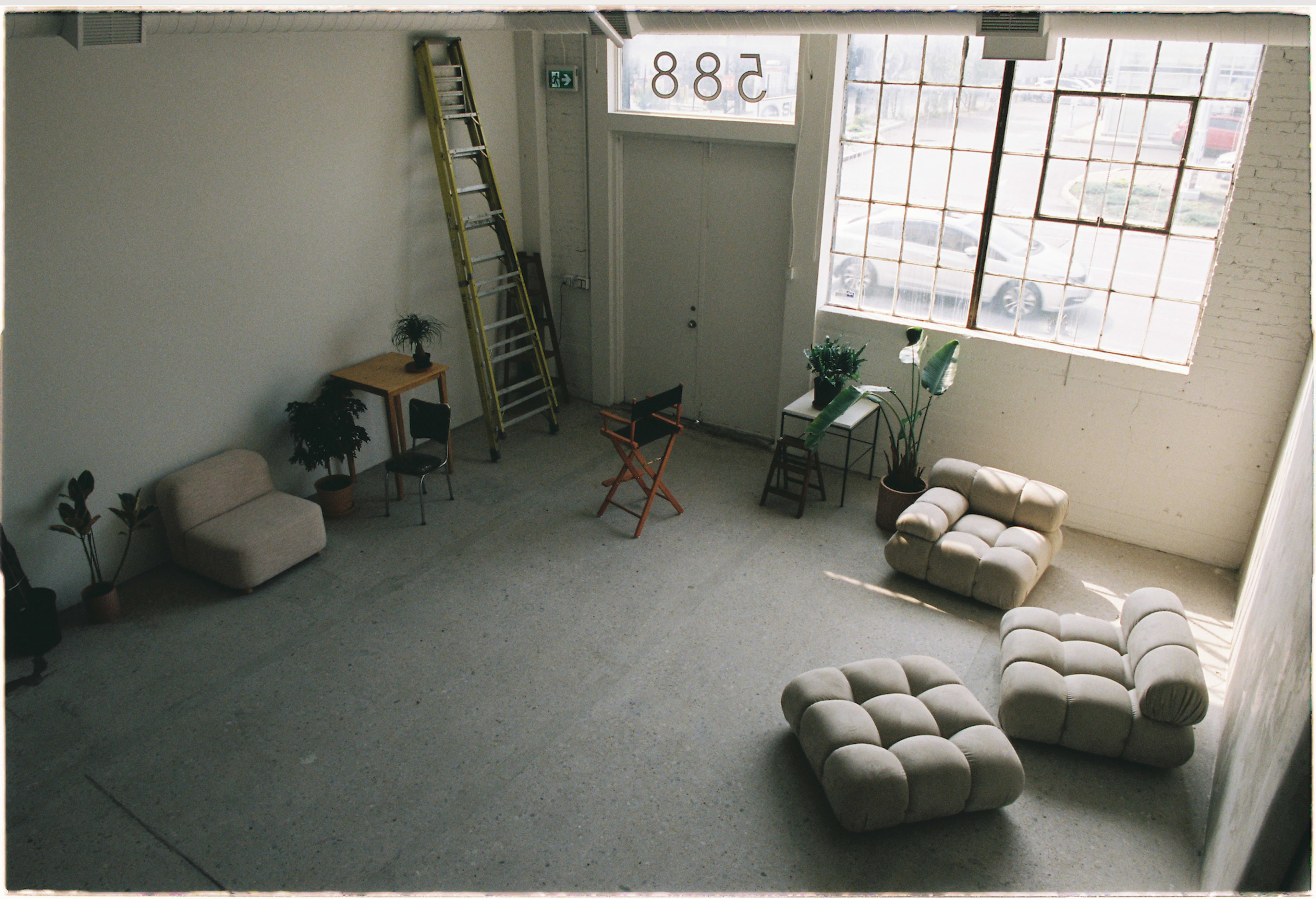 A sparsely furnished room with a tall window, a ladder, two armchairs, a table, and several plants.