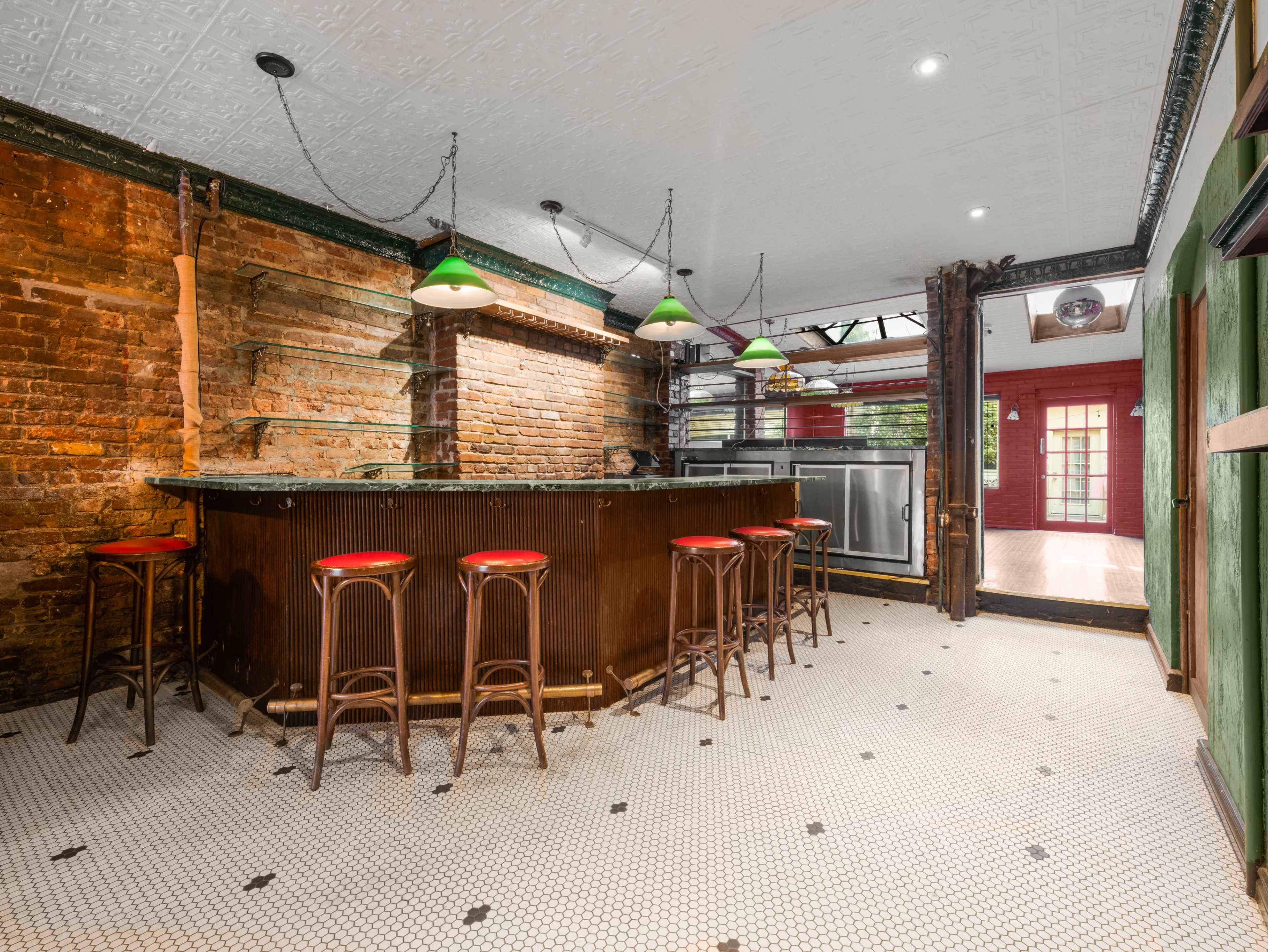 The image shows a bar area with exposed brick walls, a dark counter, red bar stools, and green pendant lights.