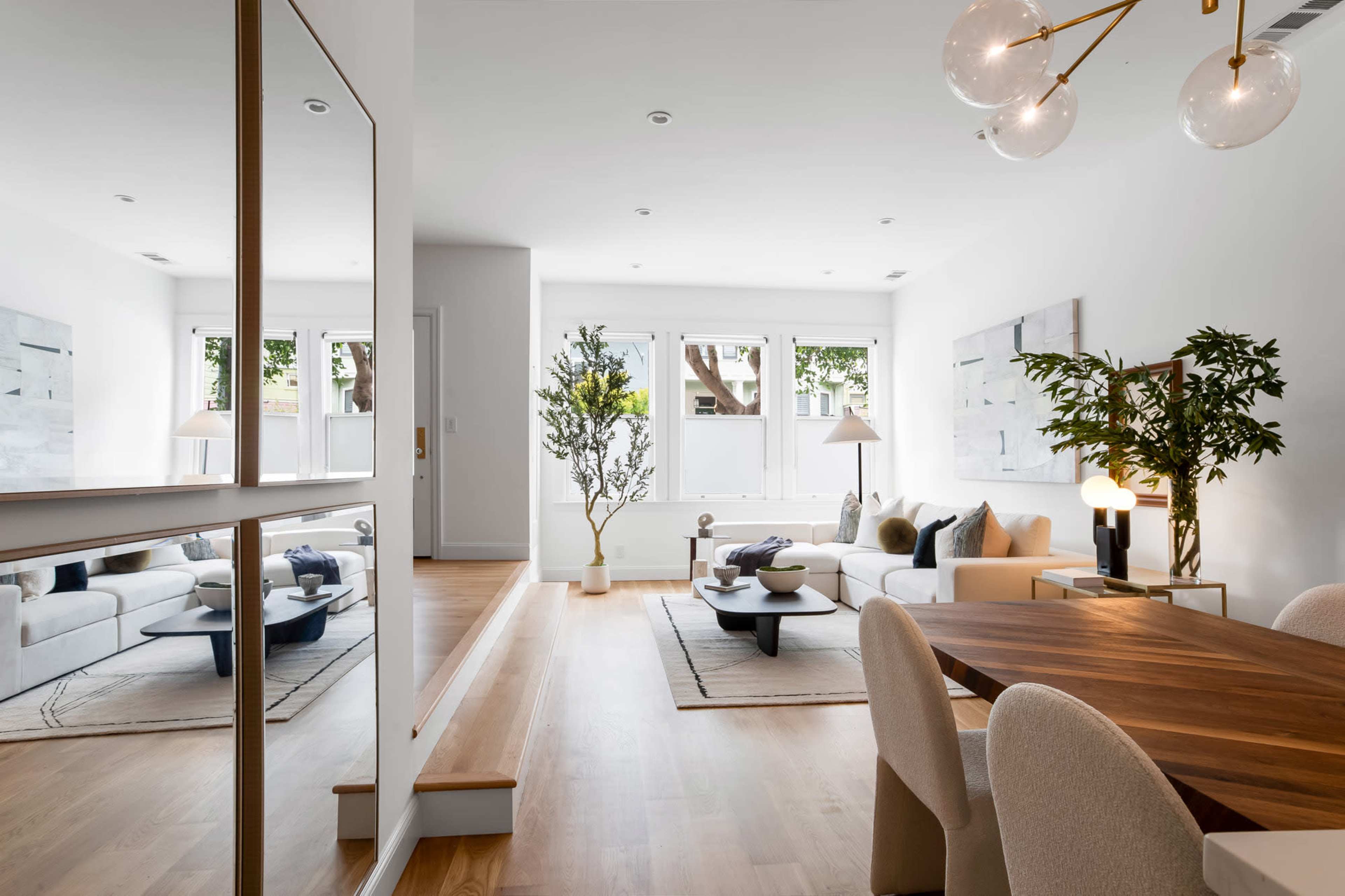 A modern living room features a white sofa, a wooden dining table, and large windows allowing natural light, with a decorative plant and minimalist decor.