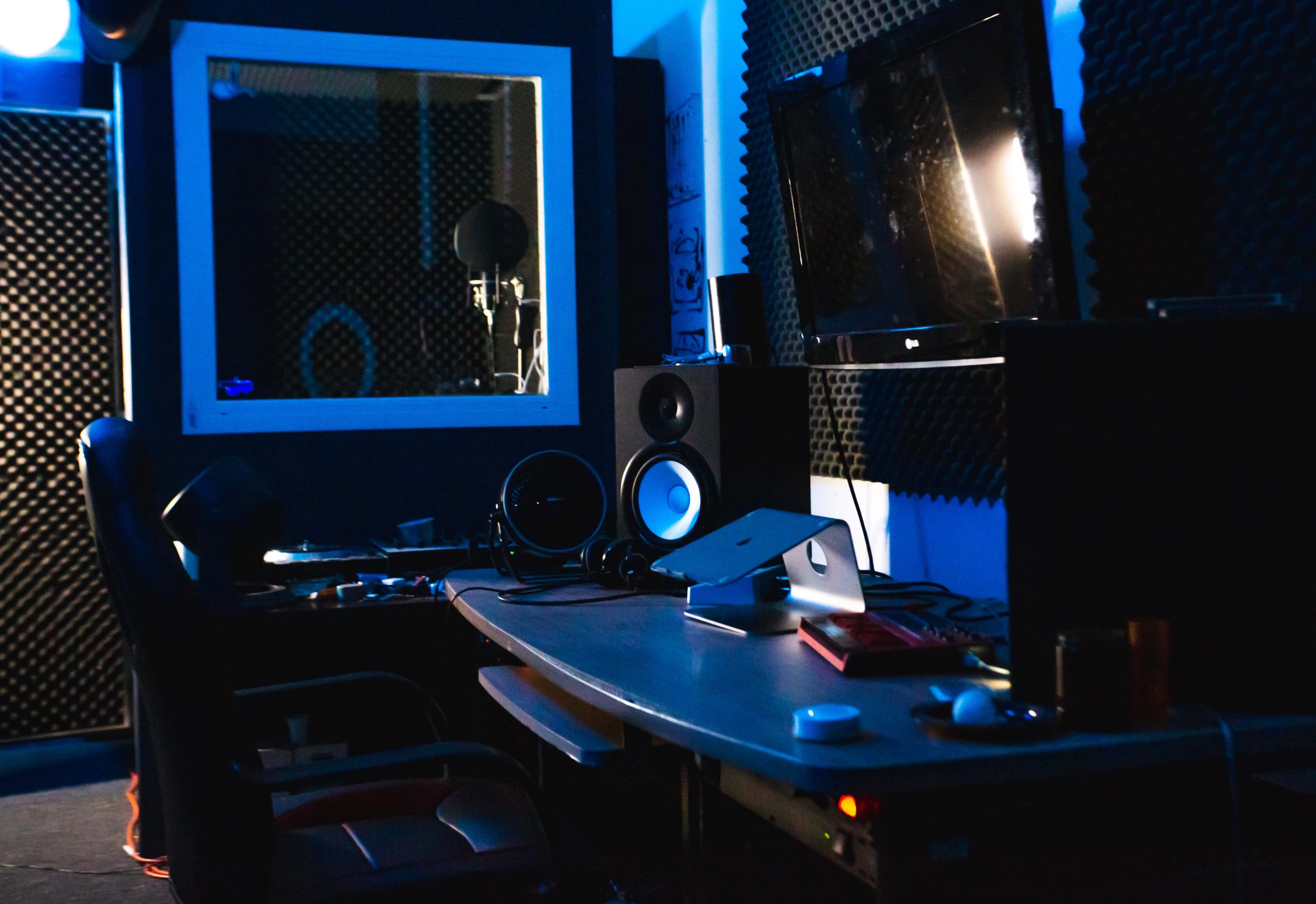 A dimly lit recording studio with soundproof walls, a desk with audio equipment, and a monitor displaying a sound setup.