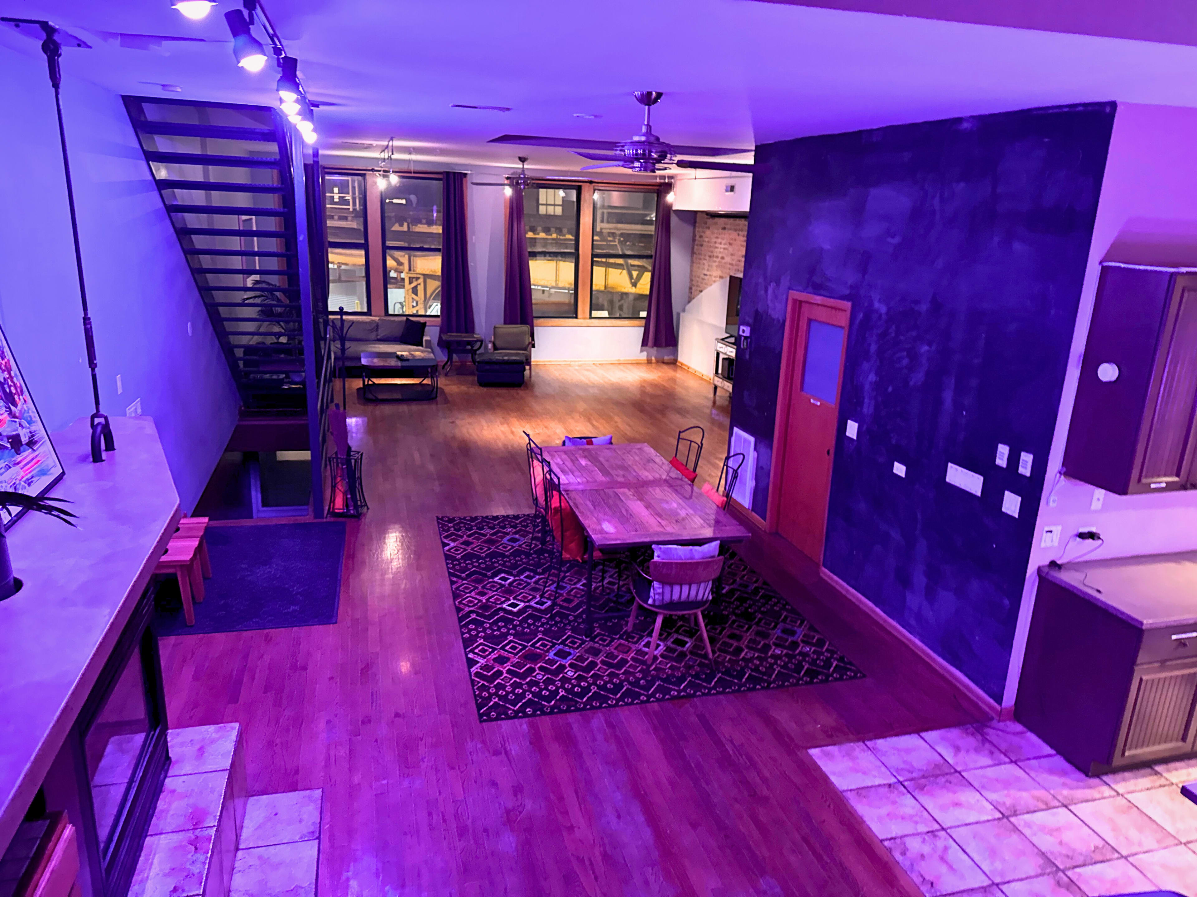 The image shows a spacious, modern loft with an open layout, featuring a dining area with a large table, a kitchen, and a living space illuminated by purple lights.