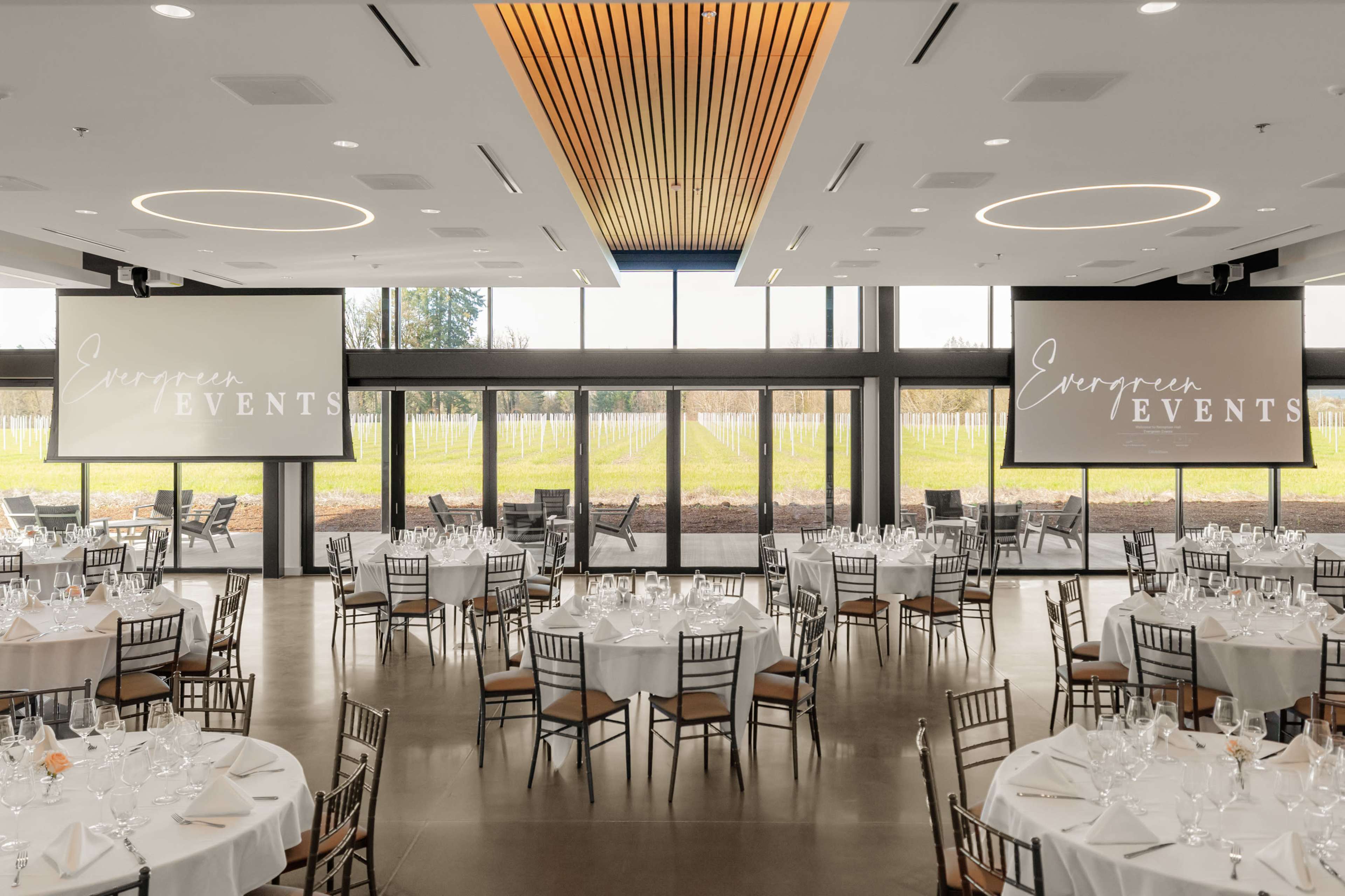 The image shows a large event space with round tables set for dining, large windows overlooking a vineyard, and screens displaying "European Events."