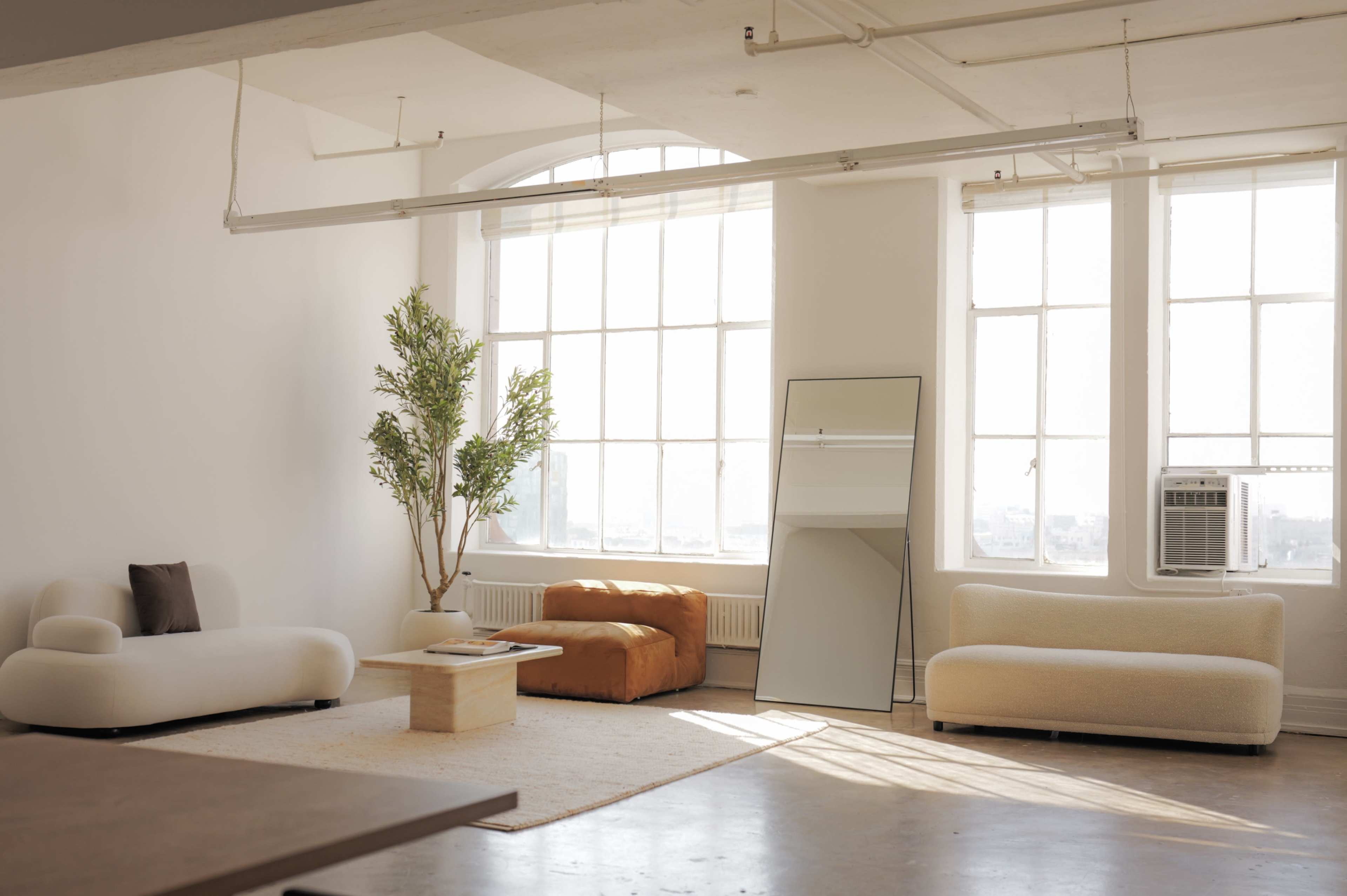 A bright, minimalist living room features large windows, a potted plant, an orange chair, a mirror, and two light-colored sofas.