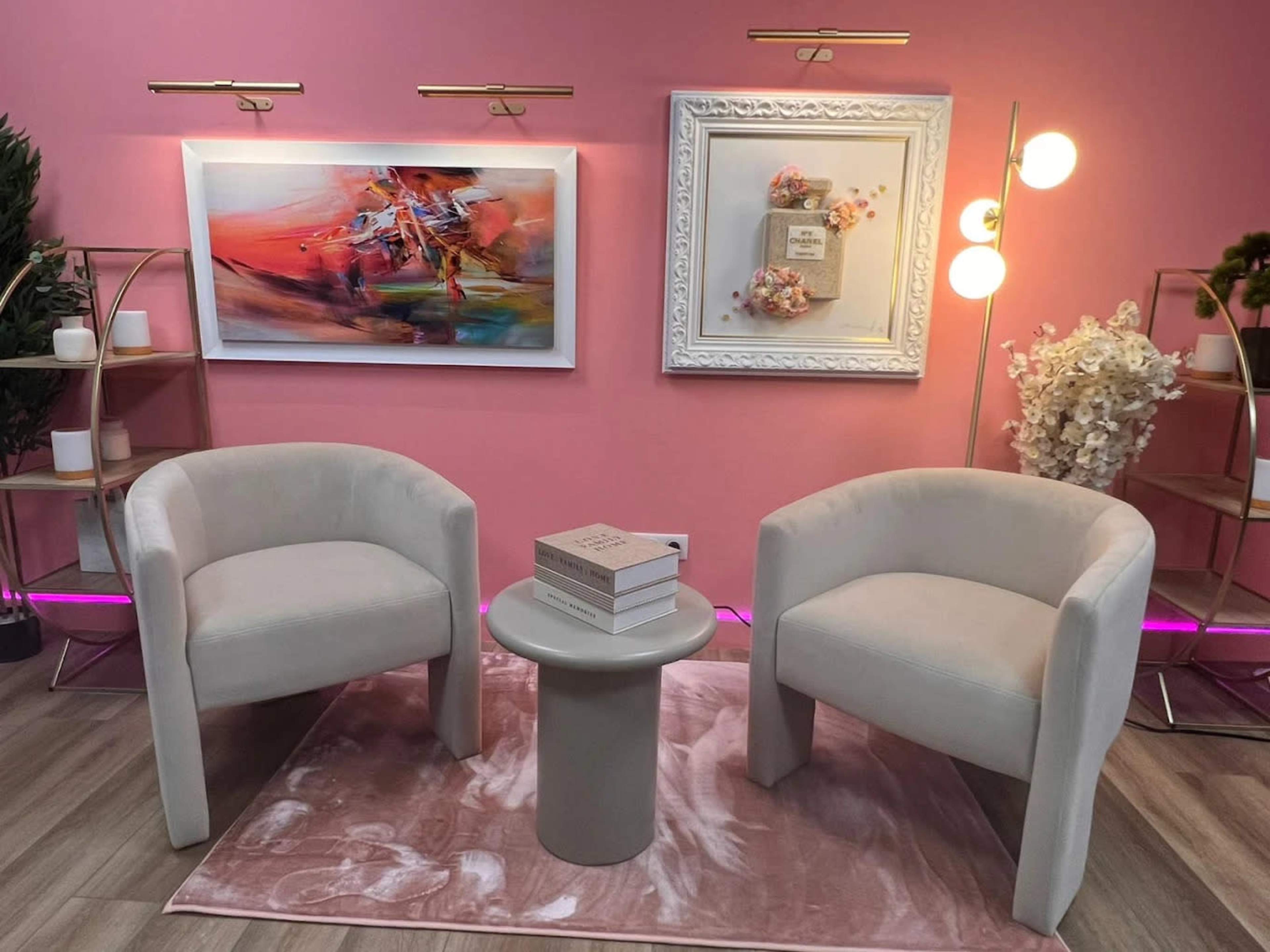 The image features two white chairs flanking a small round table on a patterned rug, with modern art pieces displayed on a pink wall behind them.