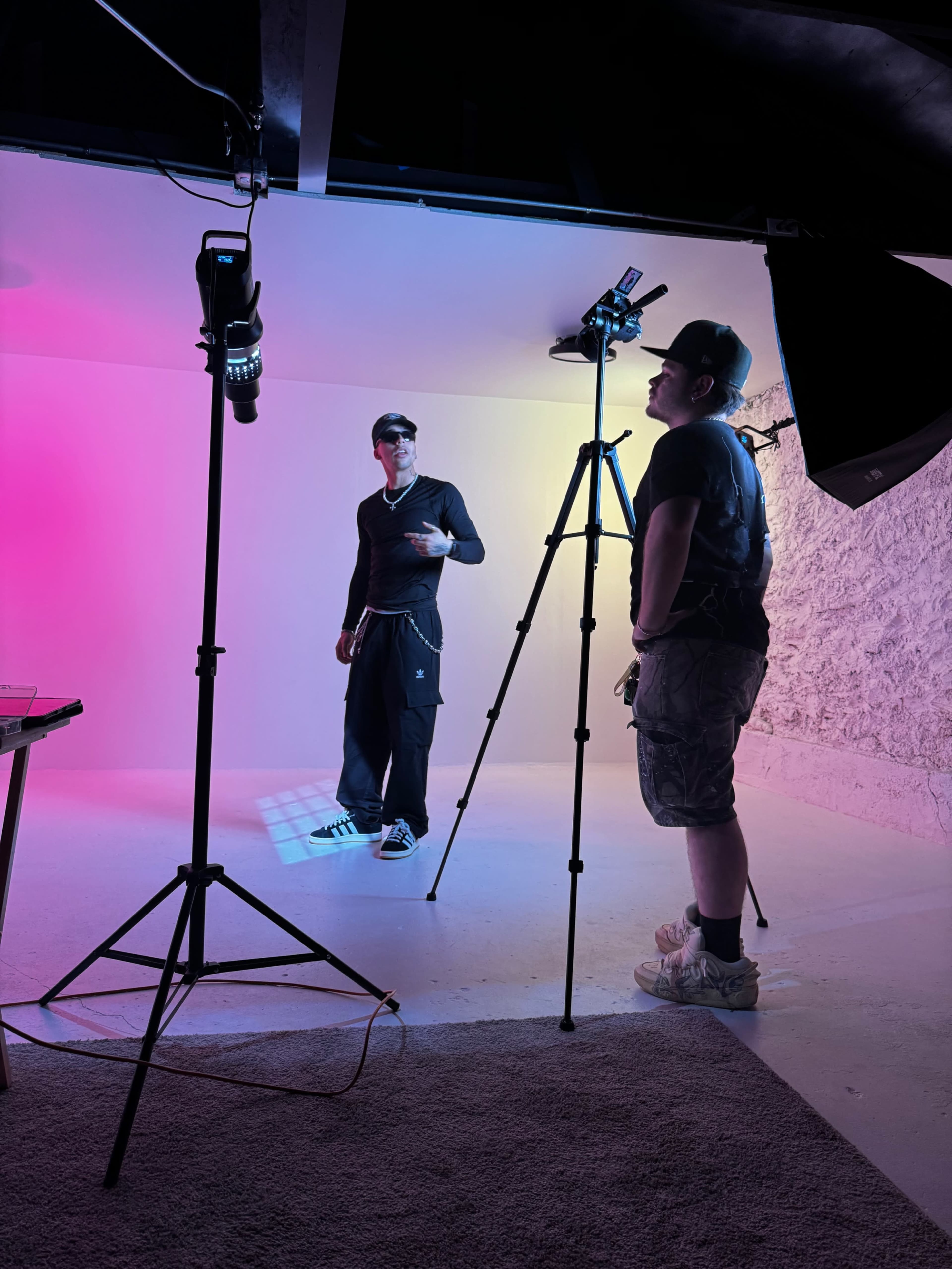 A person stands in front of a camera on a tripod, while another individual adjusts the equipment in a brightly lit studio with colored backgrounds.