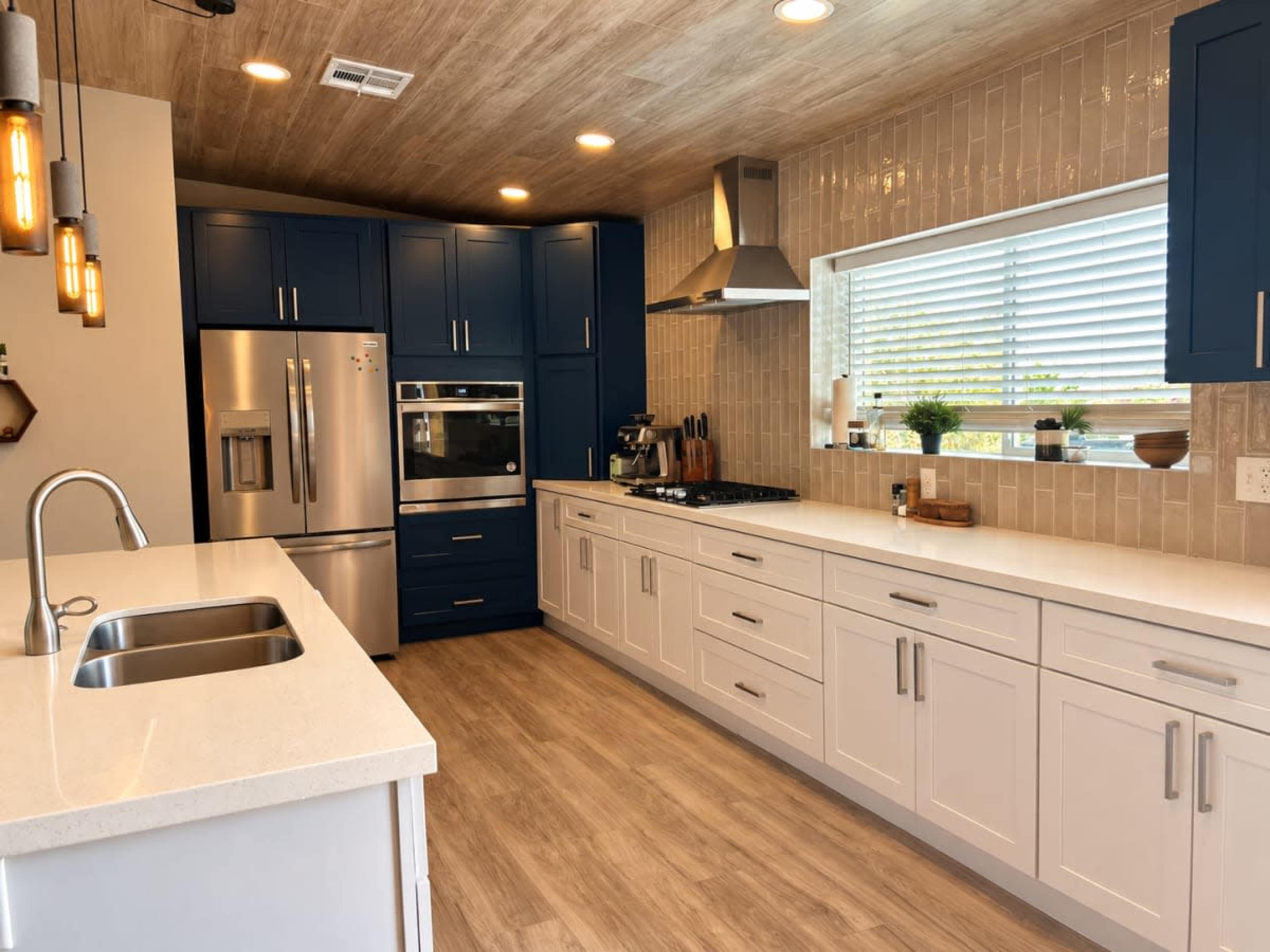 The kitchen features blue and white cabinetry, stainless steel appliances, and a light wood ceiling, with a large window allowing natural light.