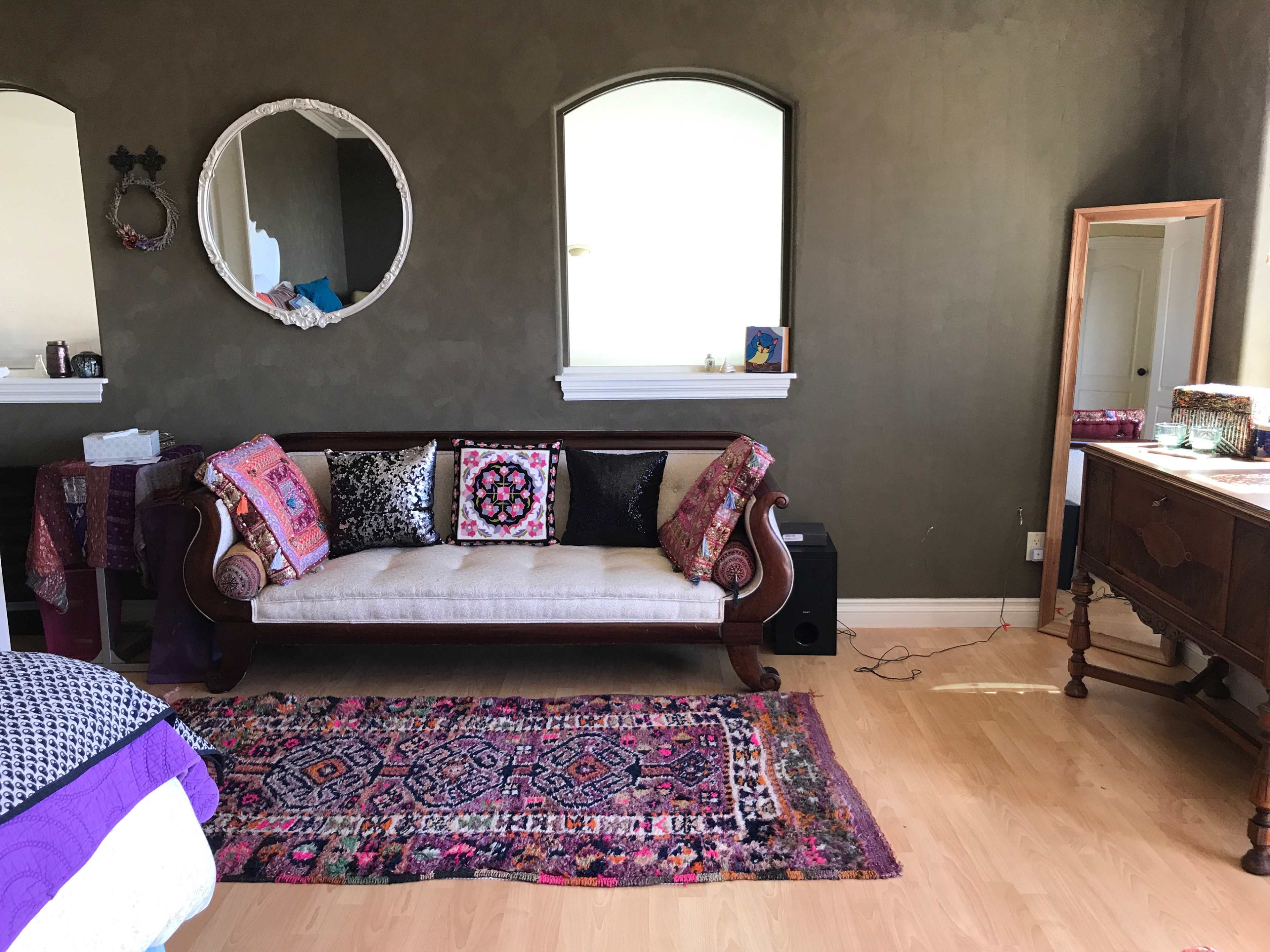 The room features a sofa with decorative pillows, a round mirror on the wall, and a vintage dresser beside a window.