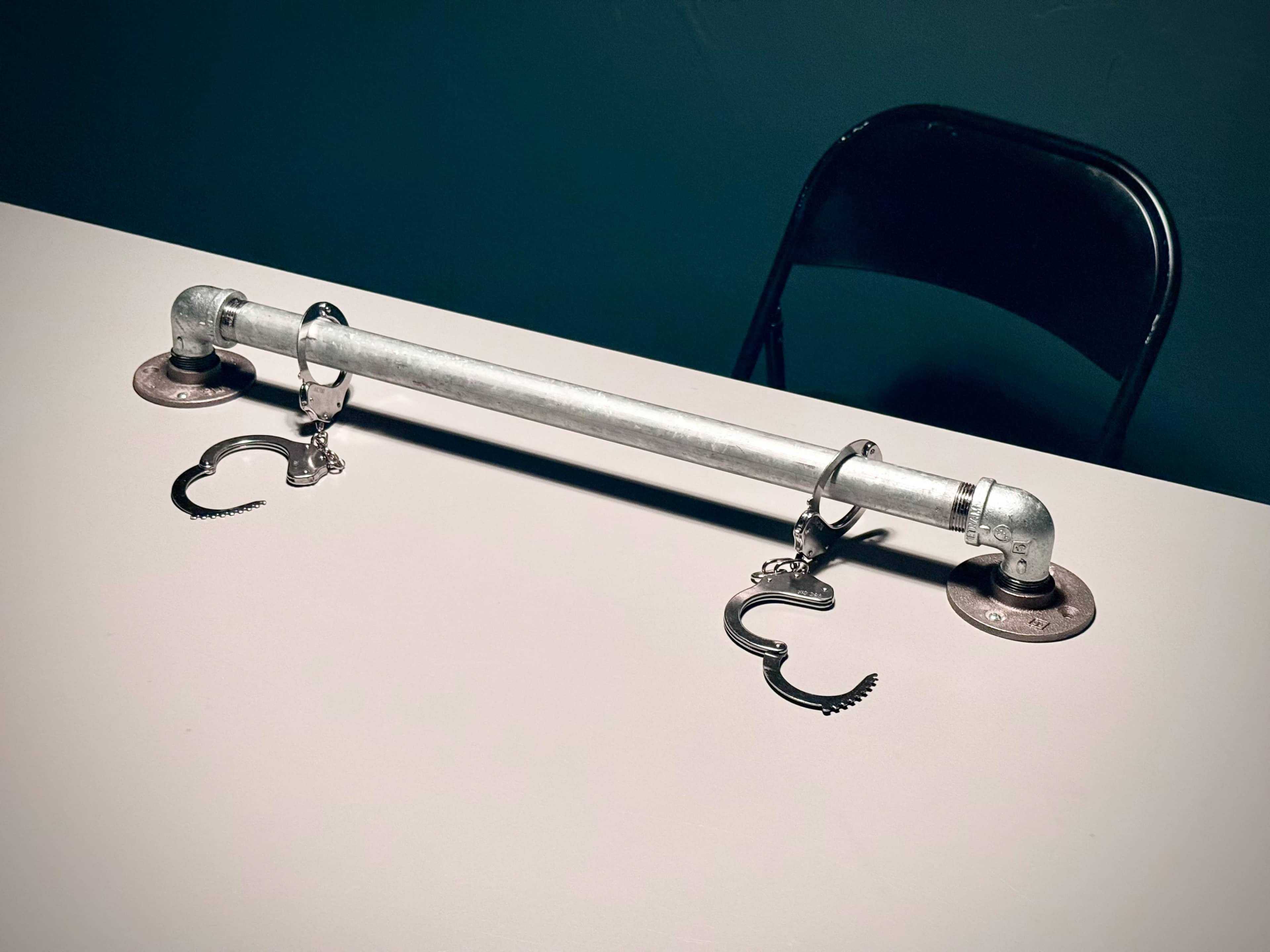 A metal pipe with handcuffs attached sits on a tabletop beside a black chair.