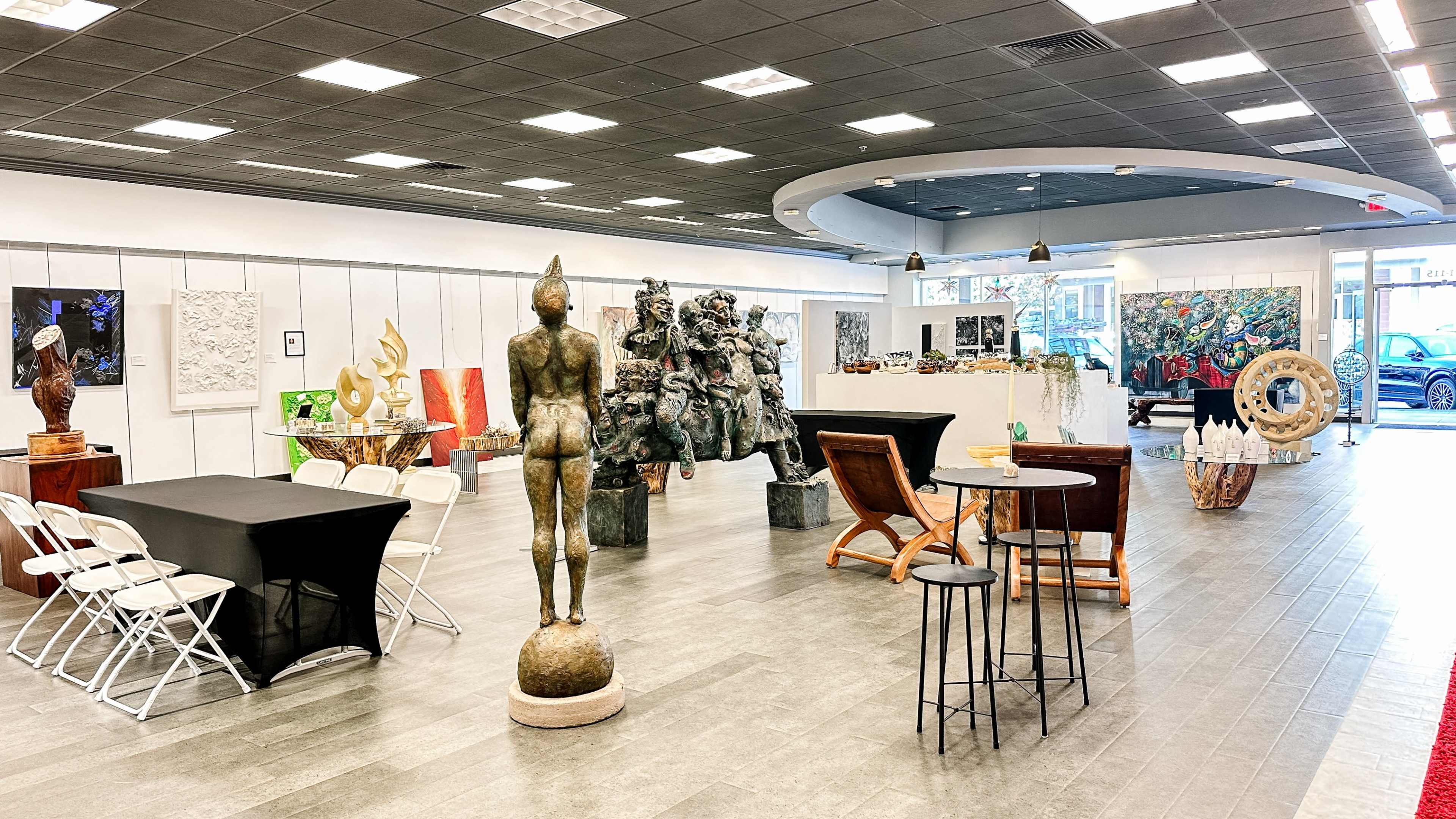 The image shows an art gallery featuring various sculptures, paintings, and seating arrangements in a spacious, well-lit interior.