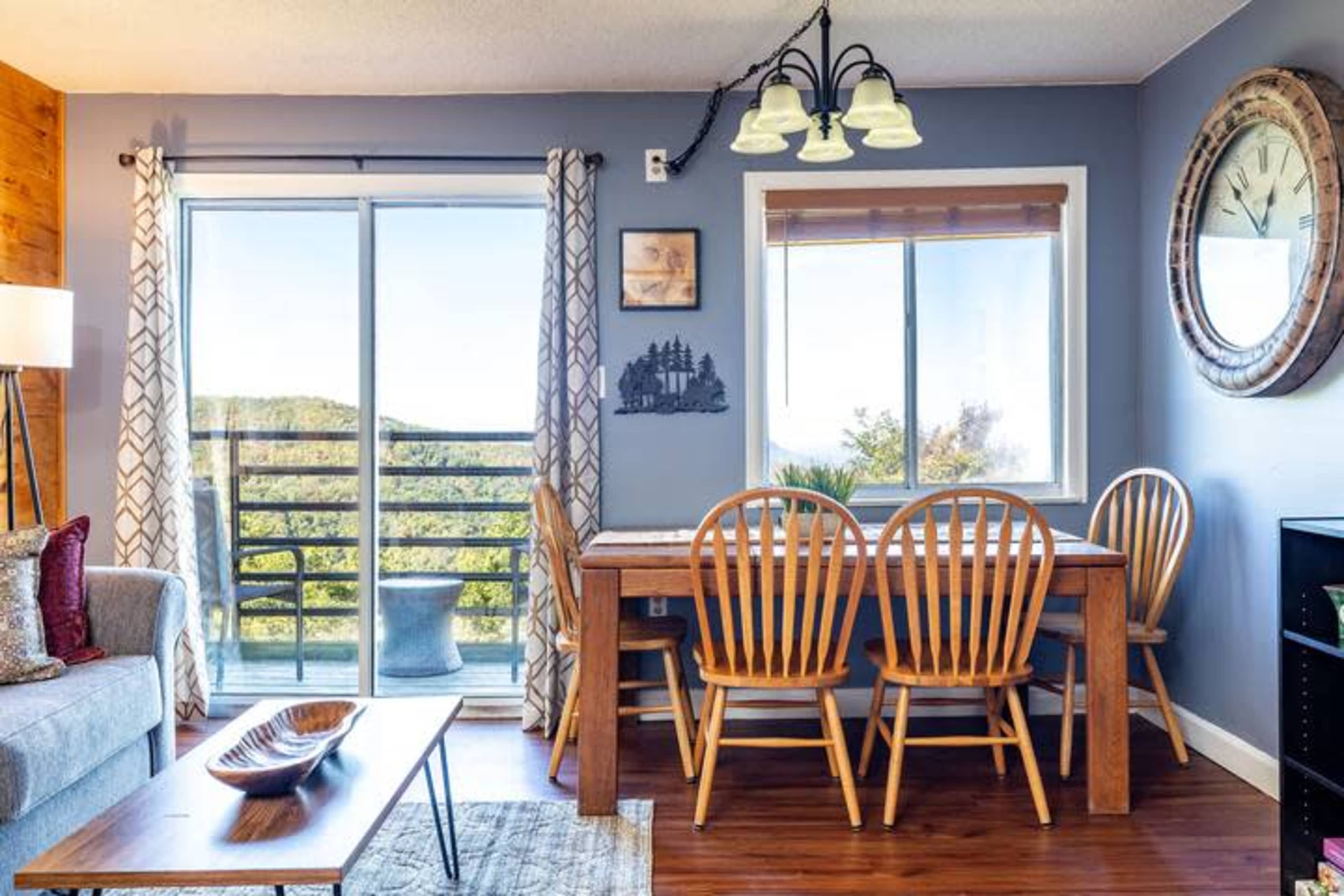 The image shows a dining area with a wooden table and chairs, a gray sofa, and large windows offering a view of the outdoor landscape.