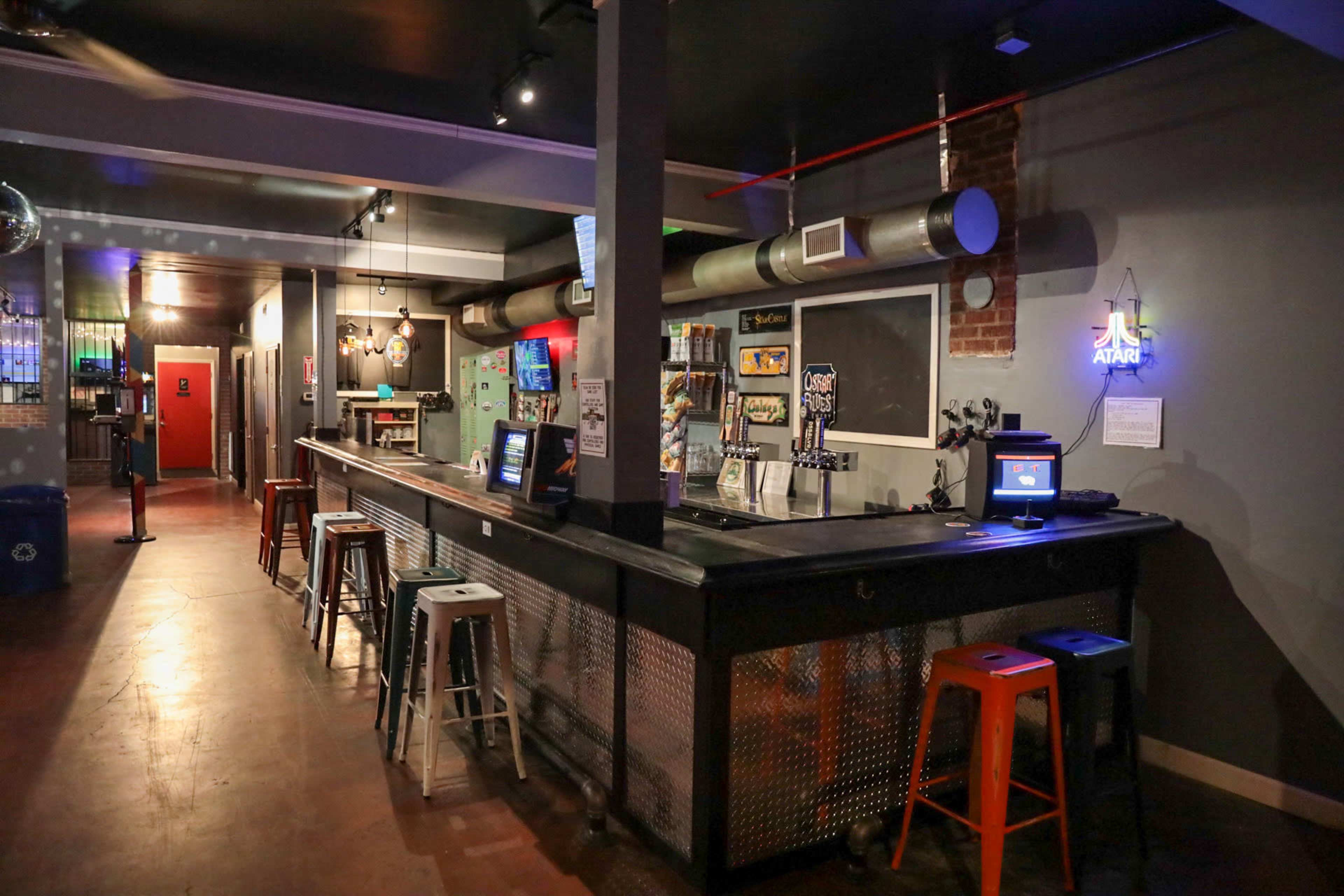 A modern bar with a long counter, high stools, and colorful decor, featuring a mix of industrial and retro elements.