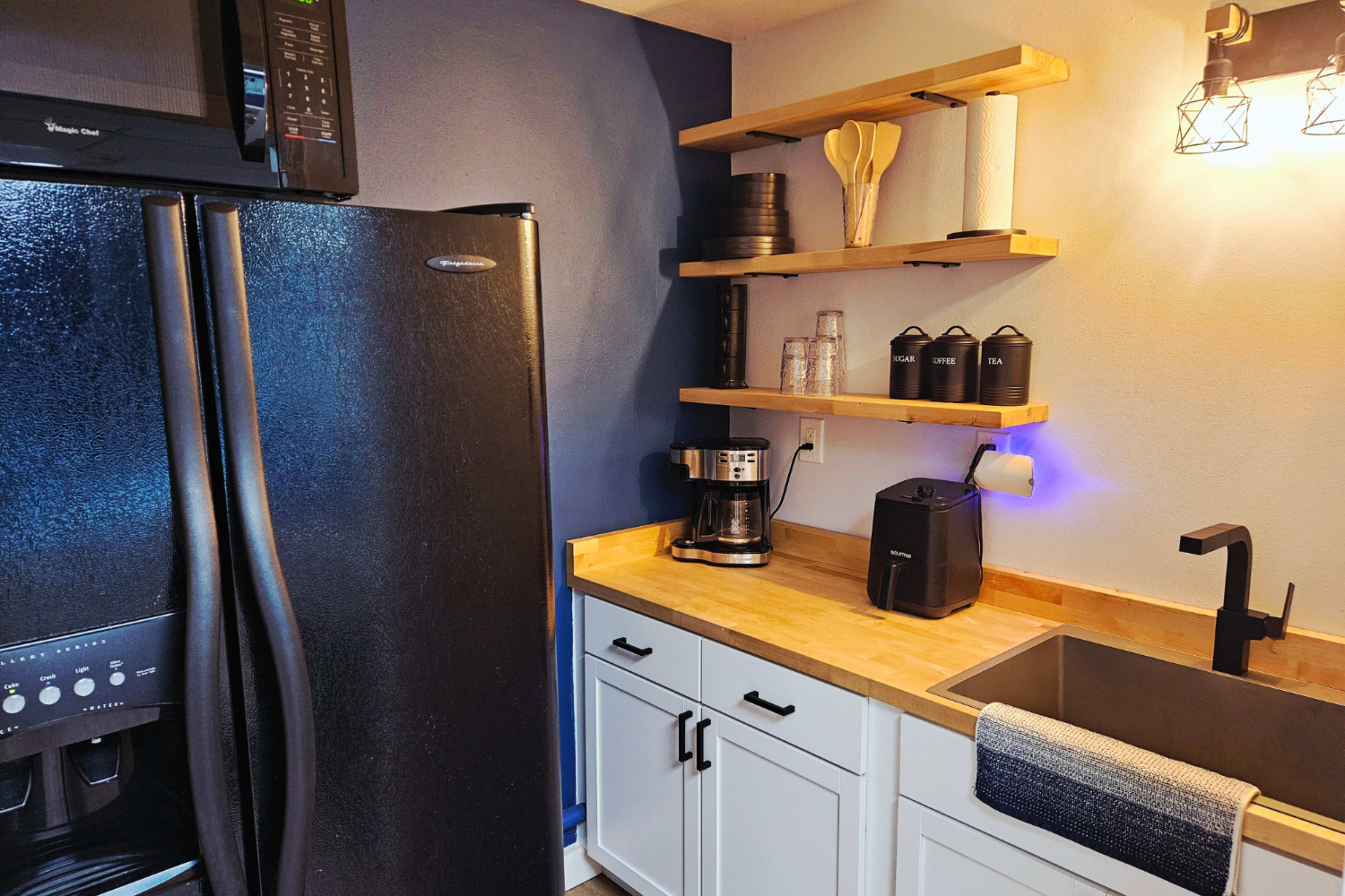 The image shows a kitchen corner featuring a black refrigerator, wooden countertops, a coffee maker, and shelves with various kitchen utensils and containers.