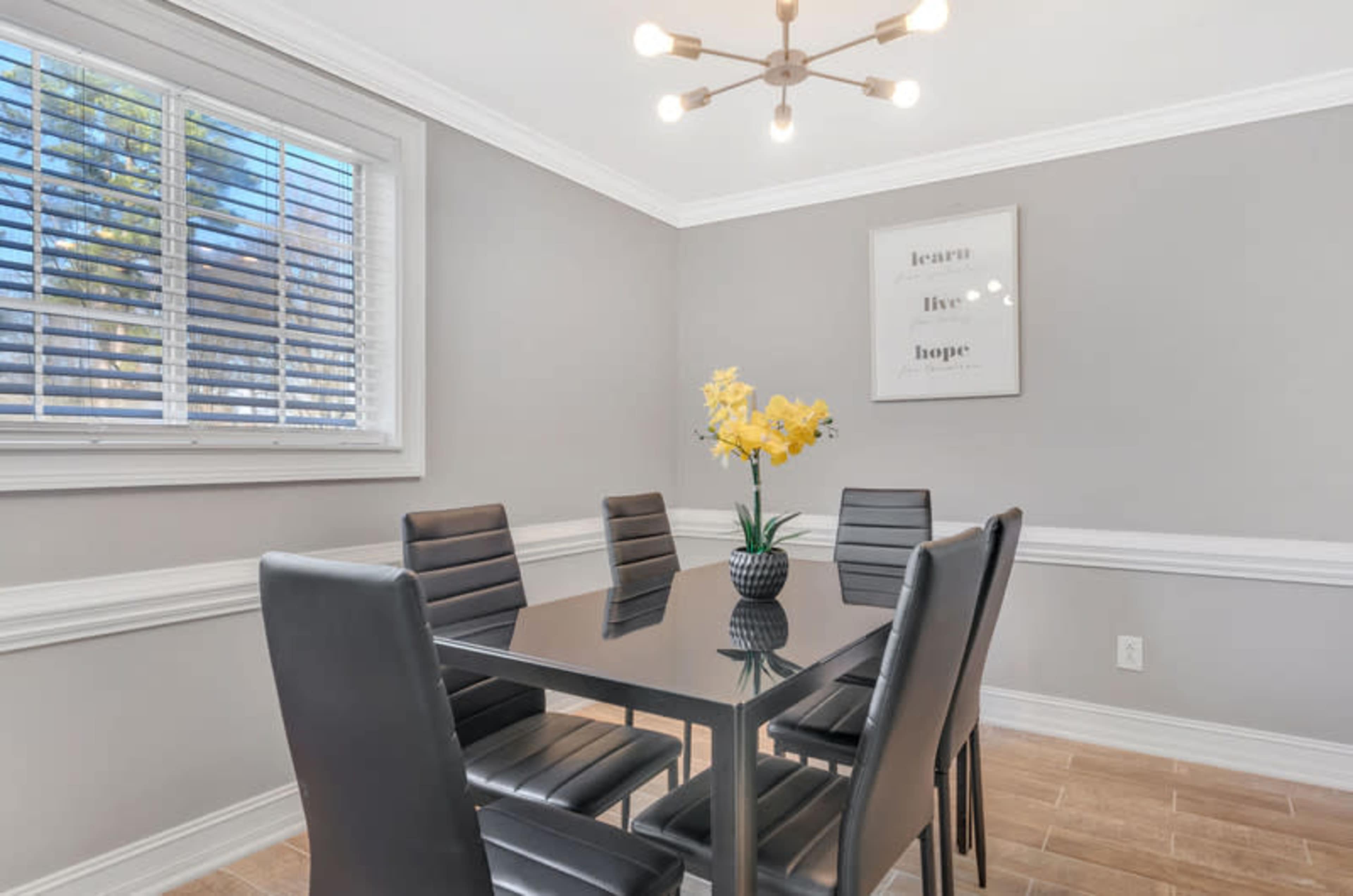 A dining area features a glass table surrounded by six black chairs, with a flower centerpiece and a framed motivational poster on the wall.