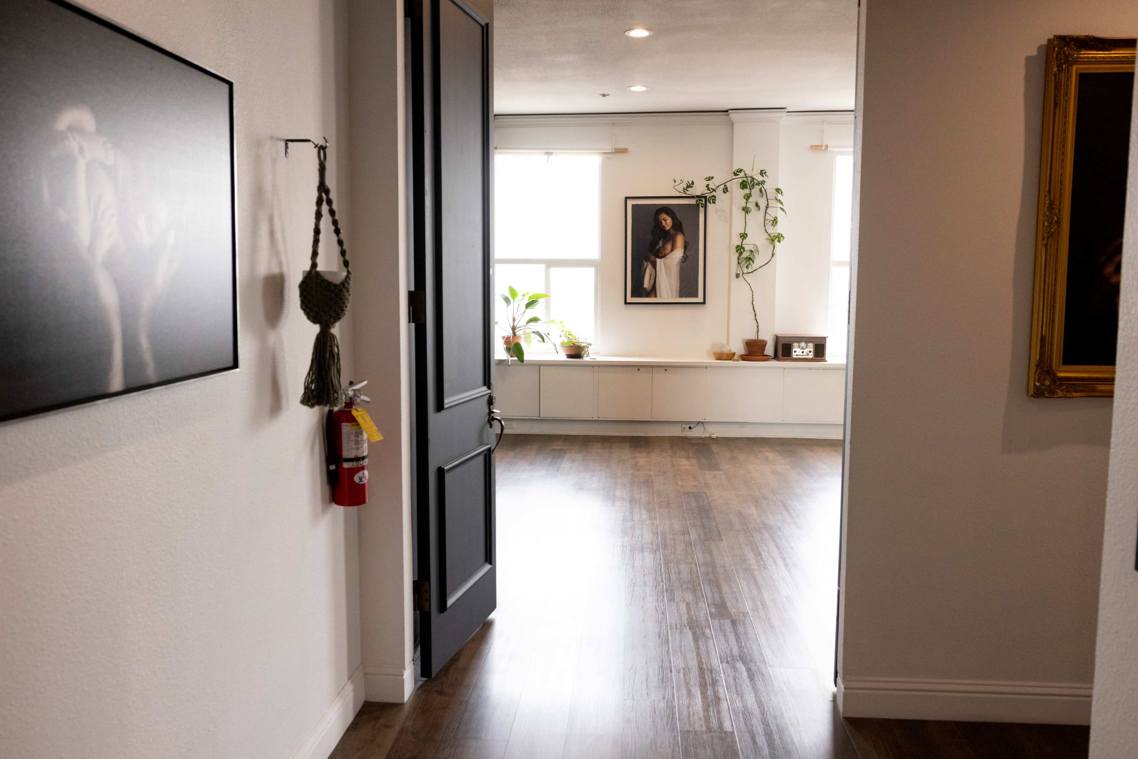 A doorway opens into a light-filled room with wooden flooring, featuring framed artwork and potted plants along the walls.