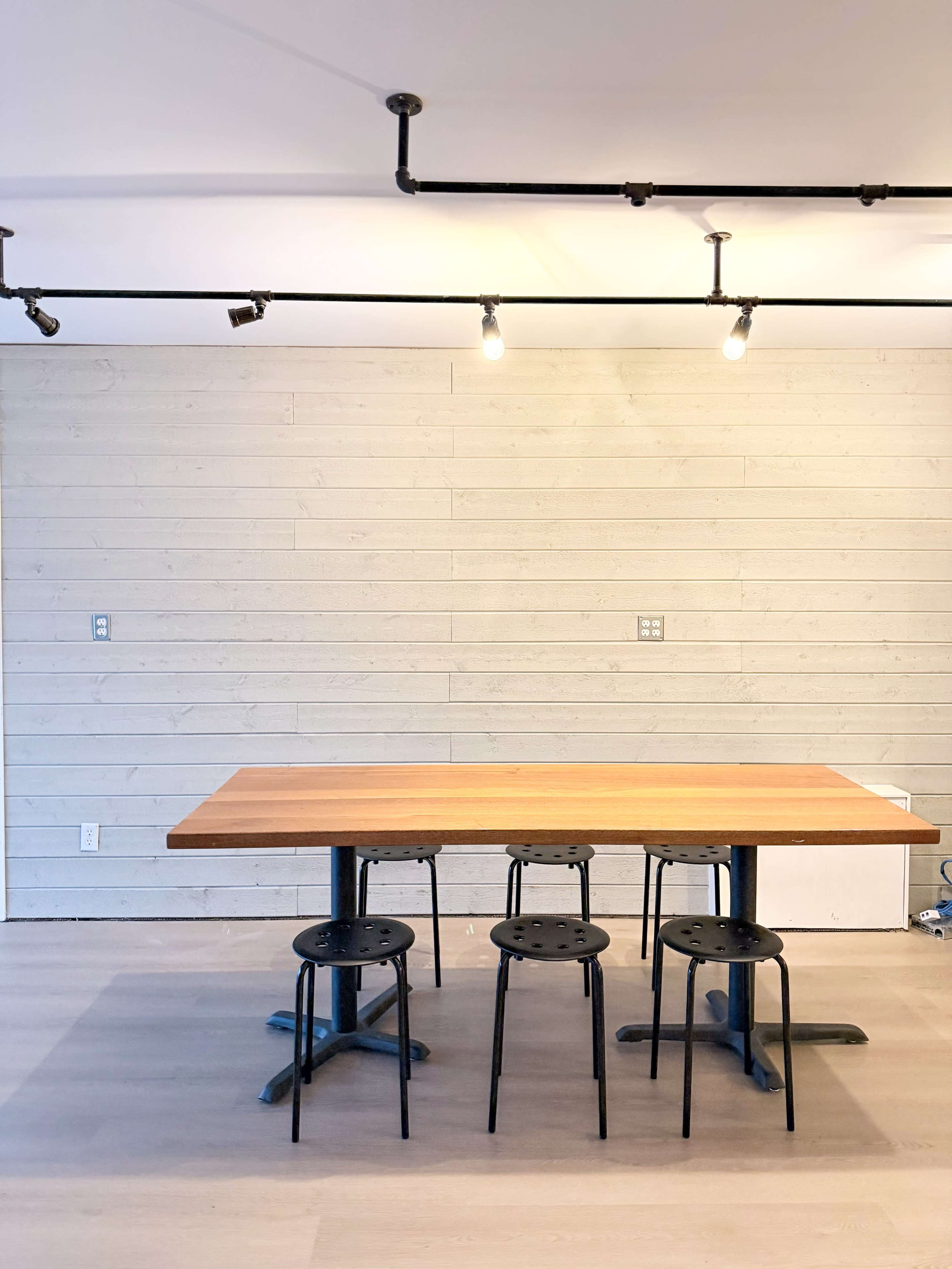 A wooden table with four black stools is positioned centrally in a room with light wooden flooring and a plain wall in the background.