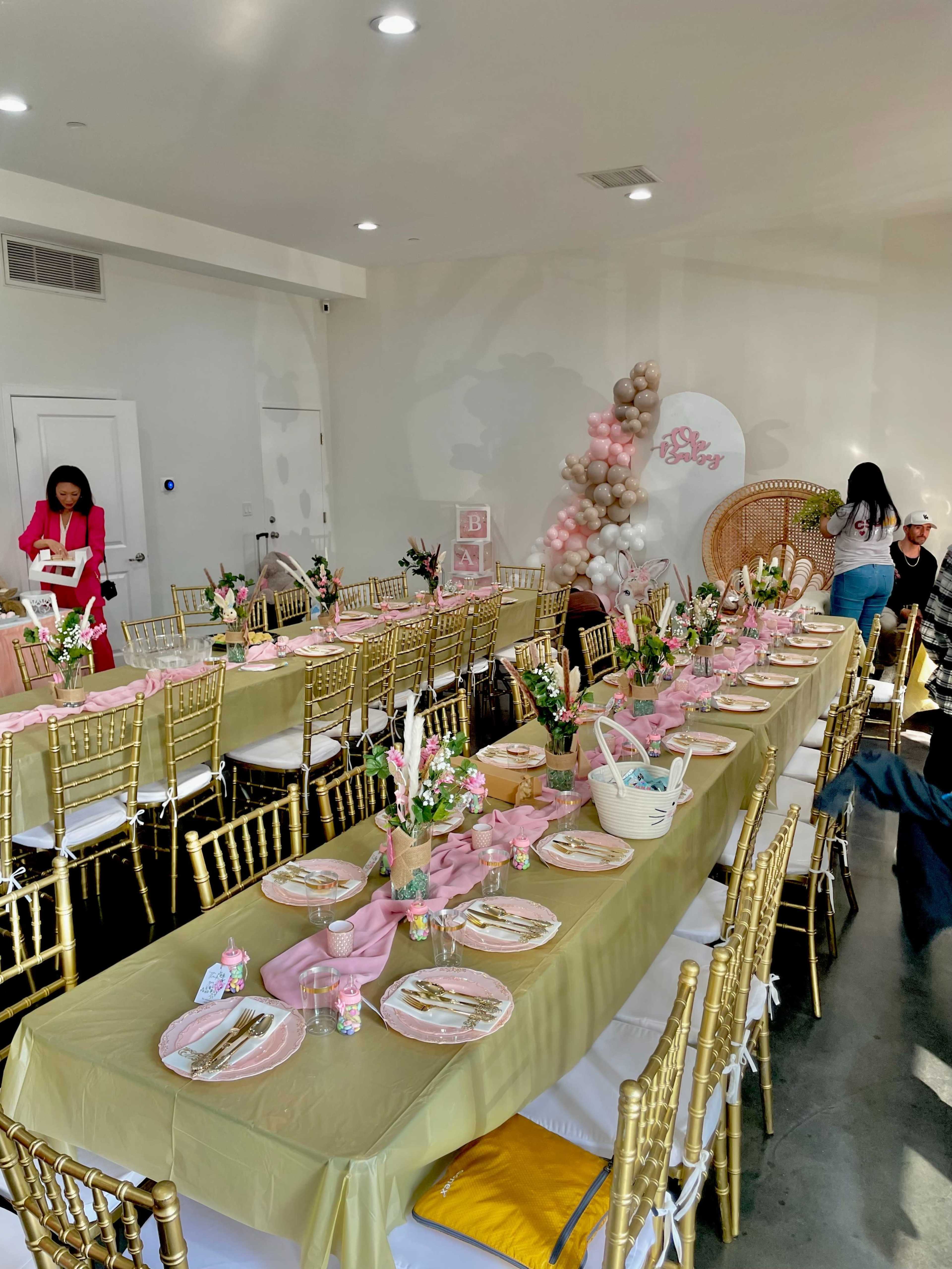 The image shows a decorated event space with a long table set for a celebration, featuring gold chairs, floral centerpieces, and pastel-colored tableware.