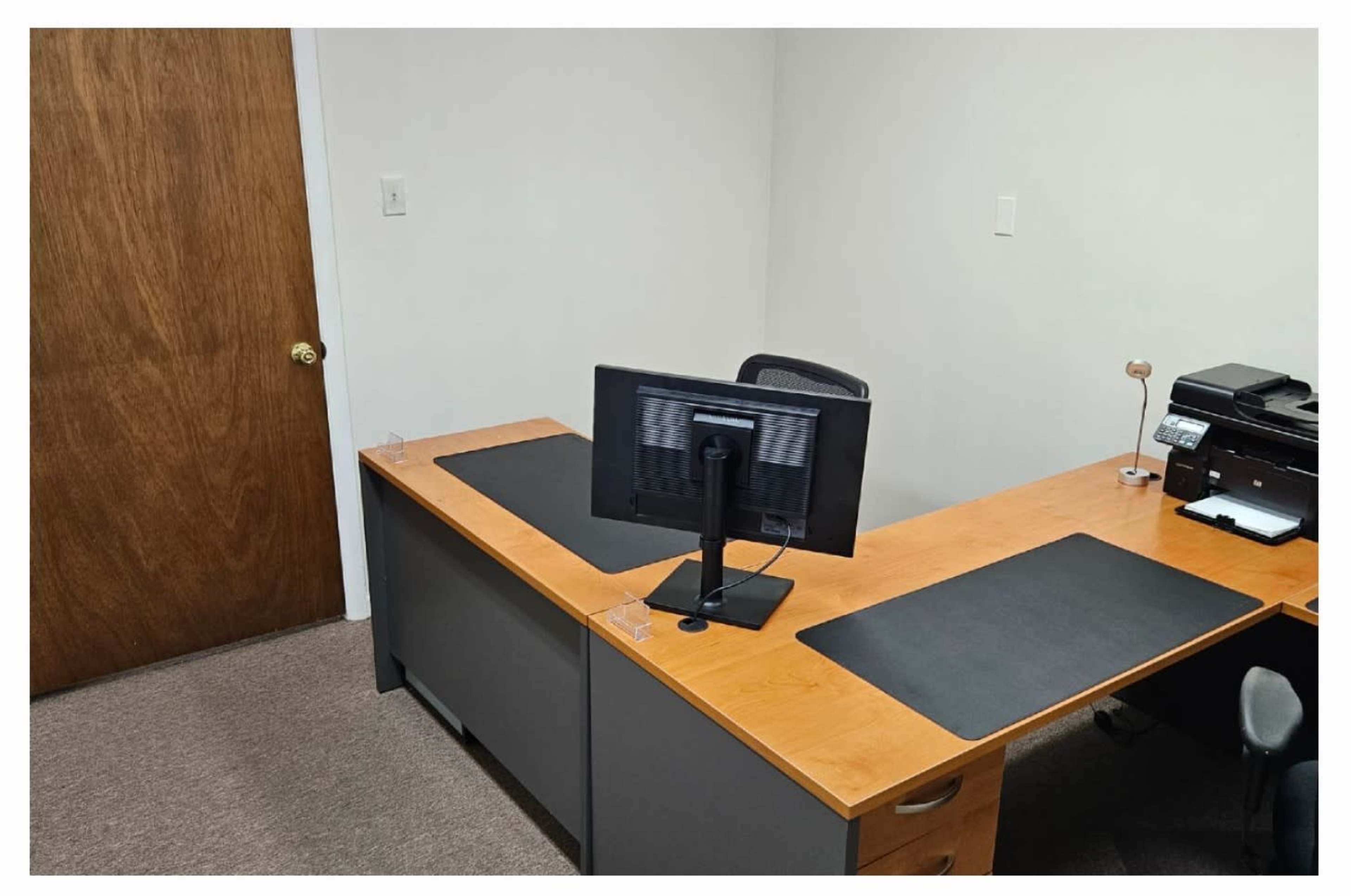 A simple office setting with a wooden desk, a computer monitor on a stand, a printer, and a closed wooden door in the background.