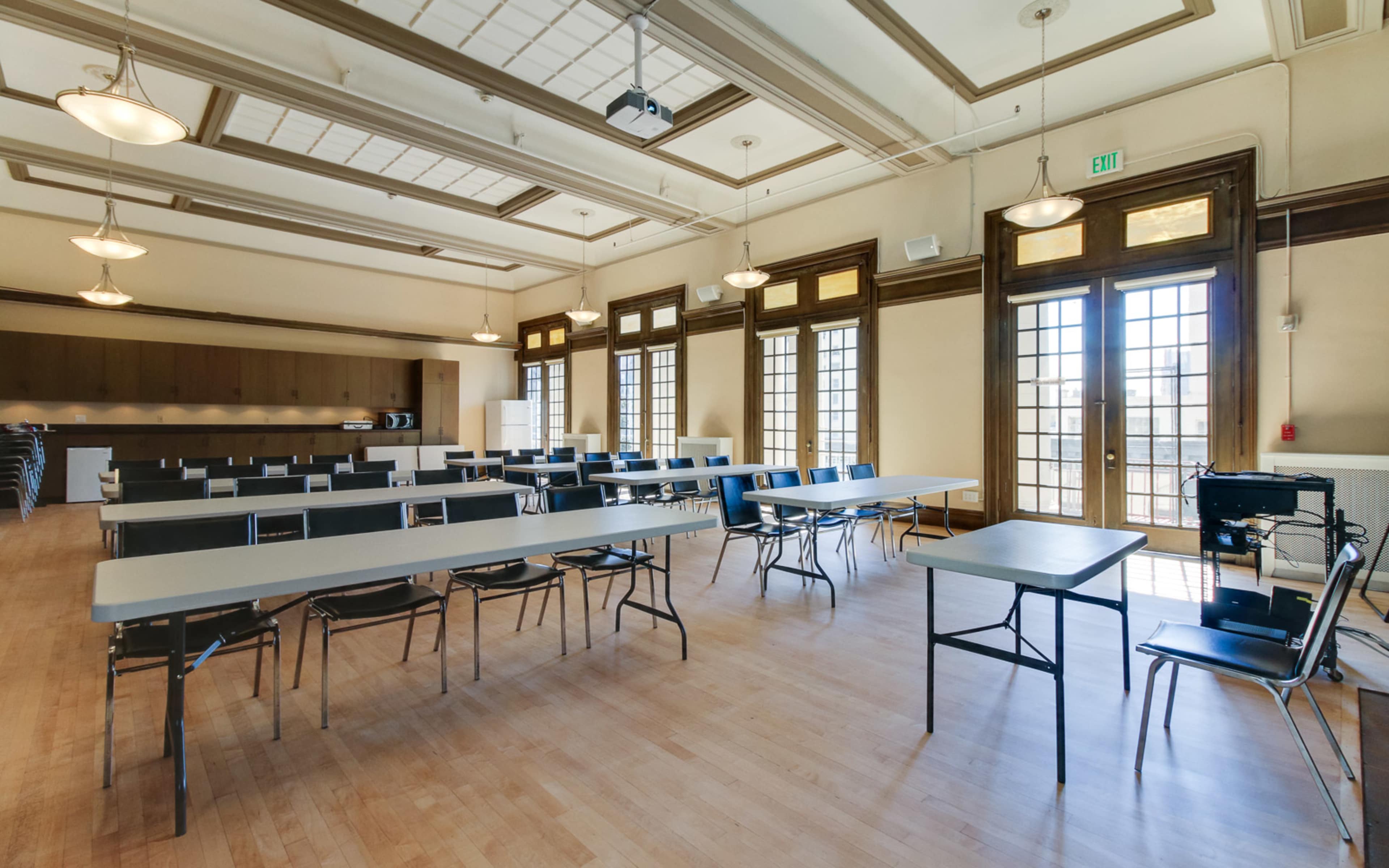 A spacious room with wooden flooring, several rows of tables and chairs arranged for seating, and large windows that allow natural light.