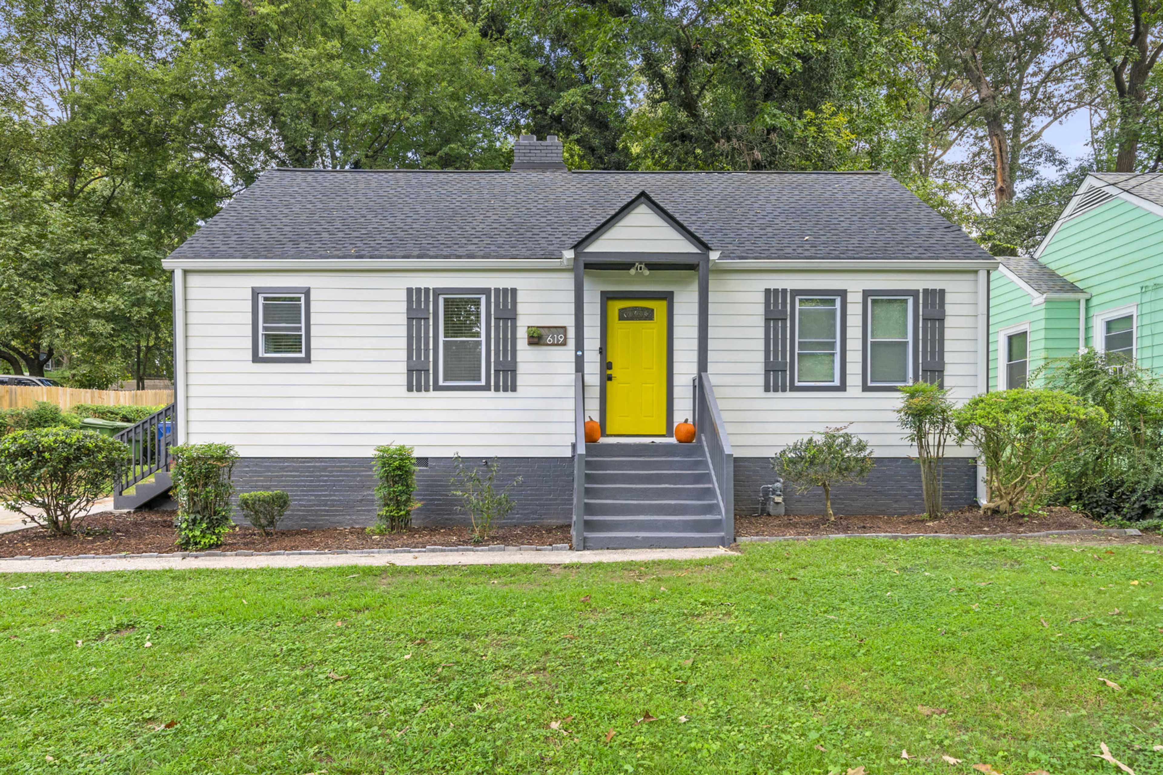 A single-story house with a yellow front door, dark shutters, and a well-maintained yard, flanked by green and white neighboring houses.