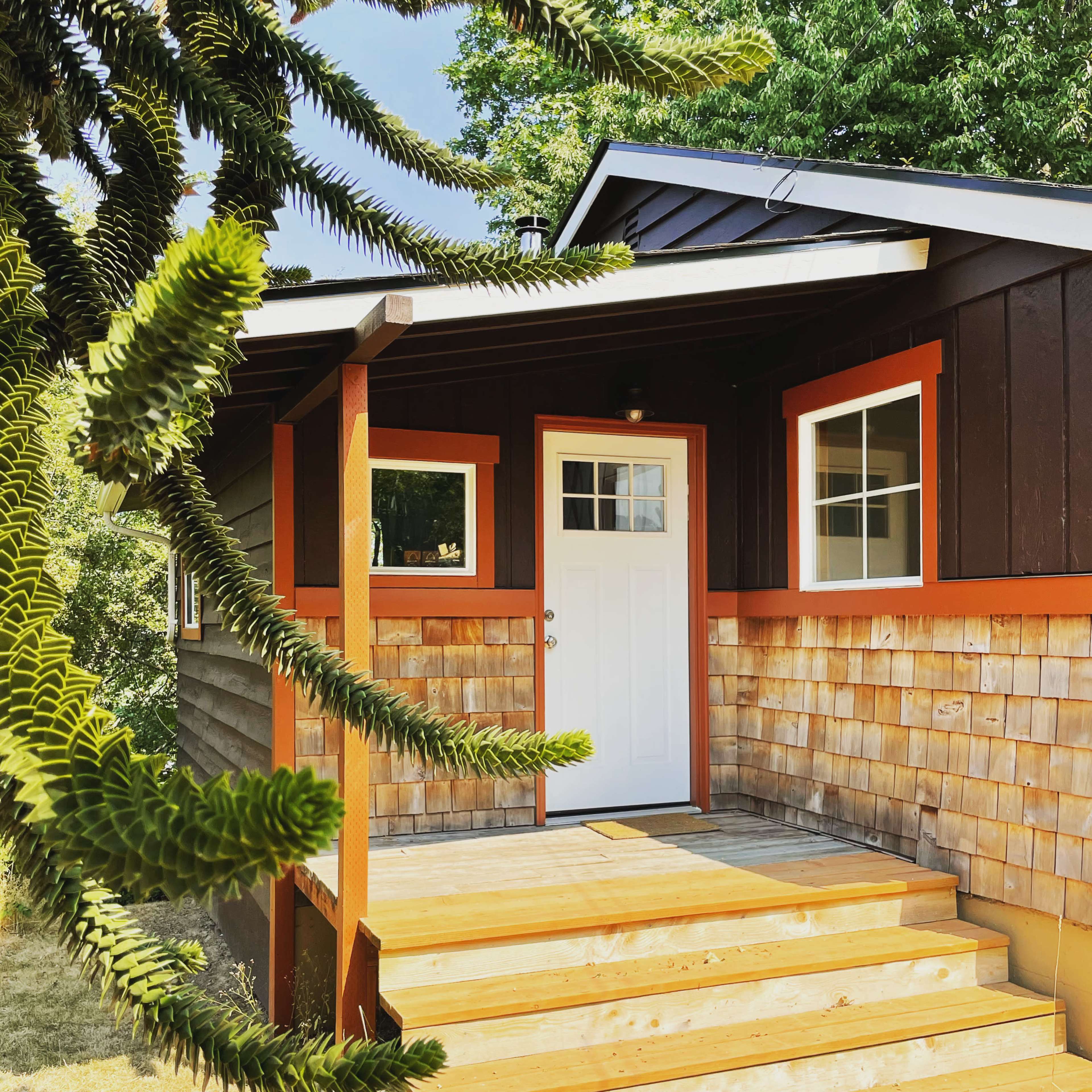 A wooden front porch leads to a dark-colored house with a white door and orange trim, surrounded by greenery.