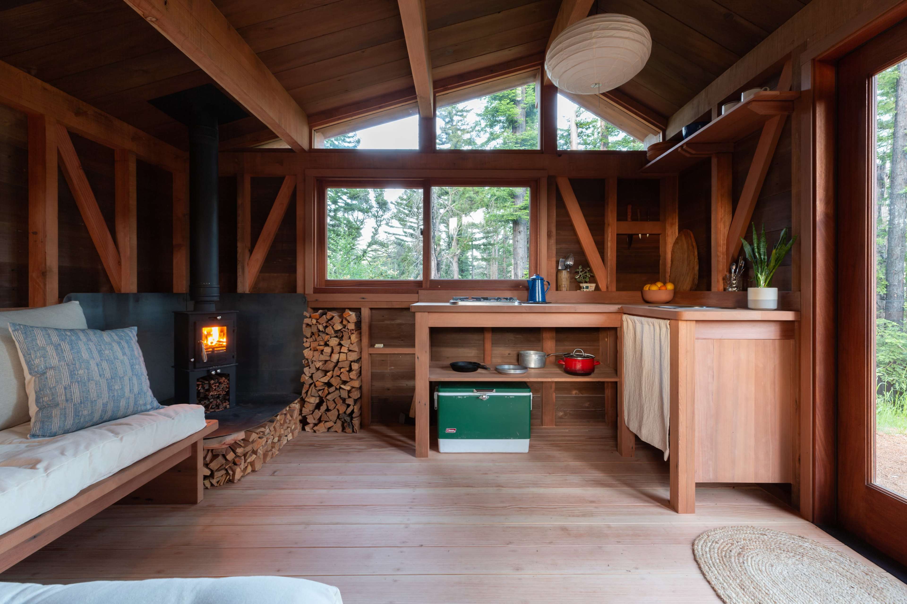 The interior of a wooden cabin features a cozy seating area, a small kitchen with wooden countertops, and a wood stove surrounded by stacked firewood.