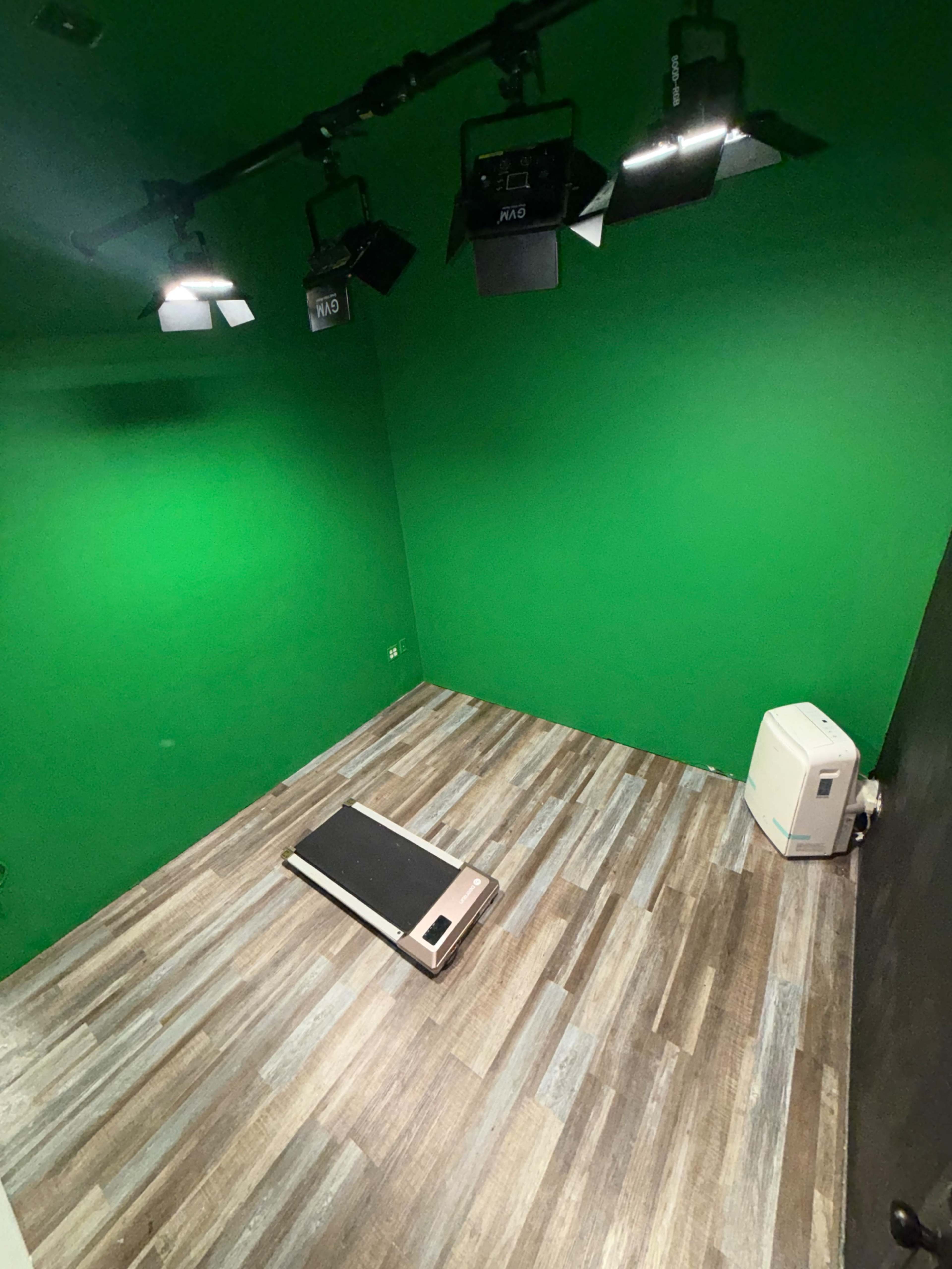 The image shows an empty room with a green wall, wooden flooring, a small treadmill on the floor, and a portable air conditioning unit.
