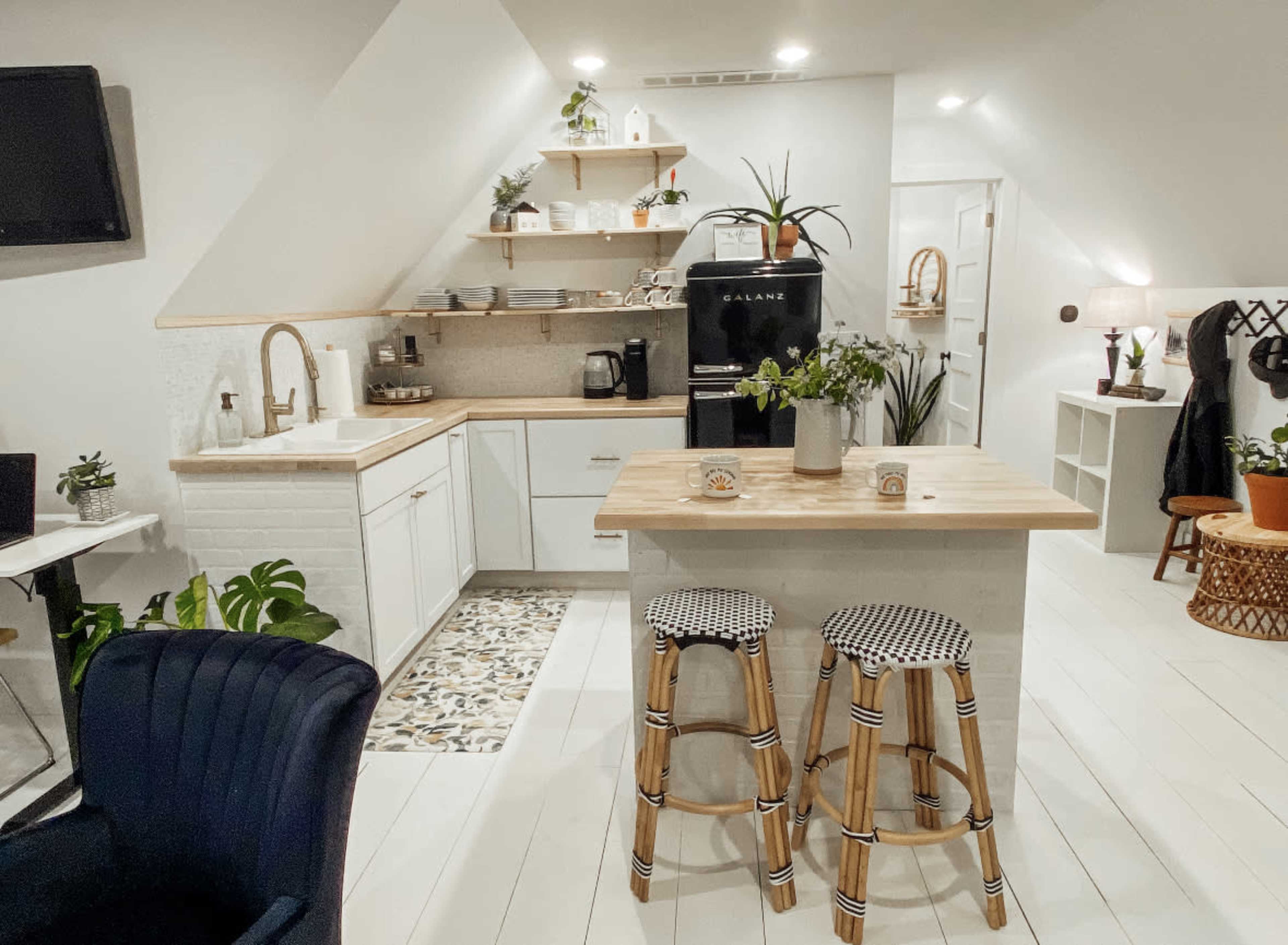 A modern kitchen features white cabinetry, a wooden countertop with two stools, and various plants arranged throughout the space.