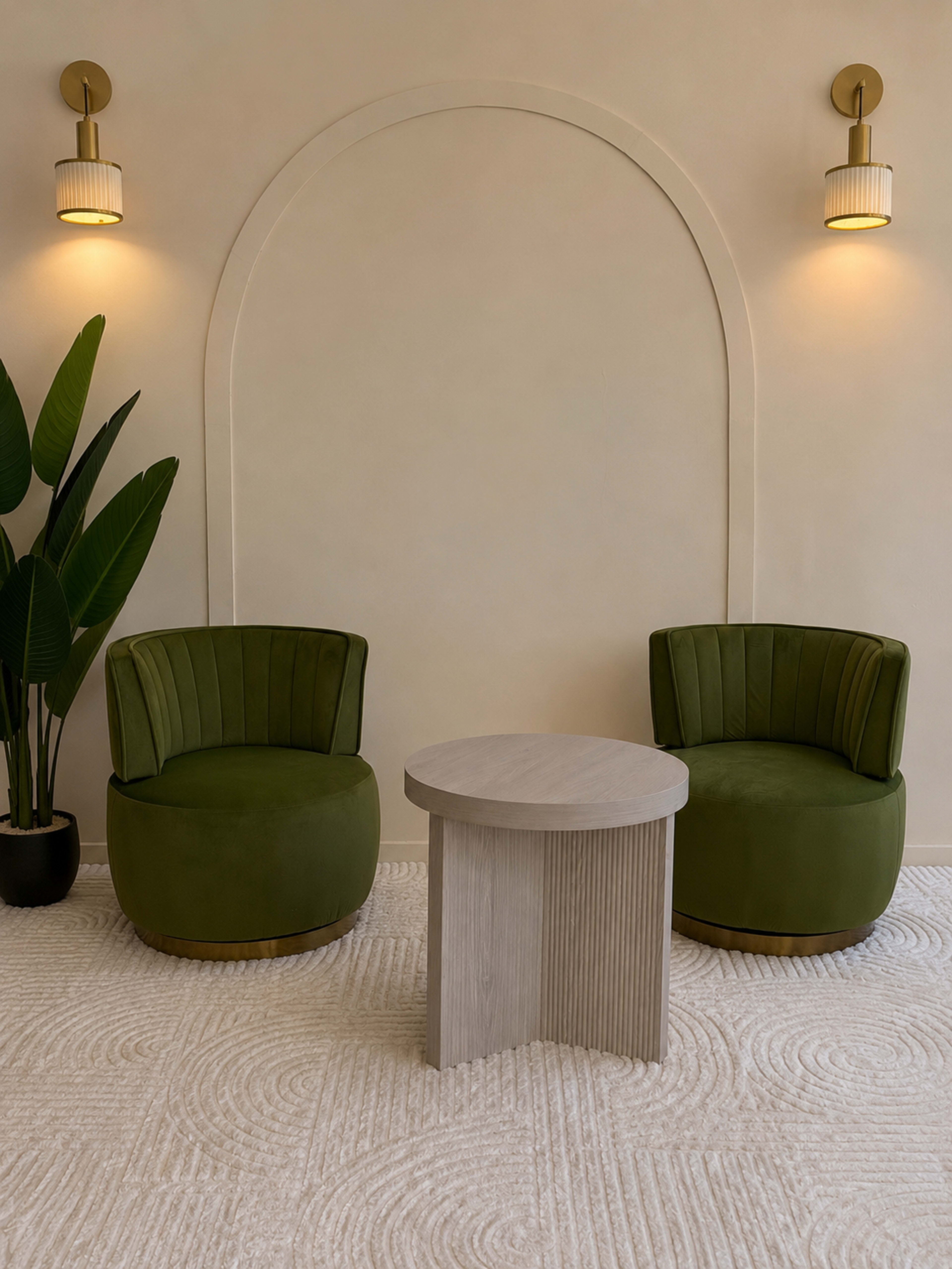The image features two green upholstered chairs flanking a circular wooden table, set against a light-colored wall with an arch and sconces.