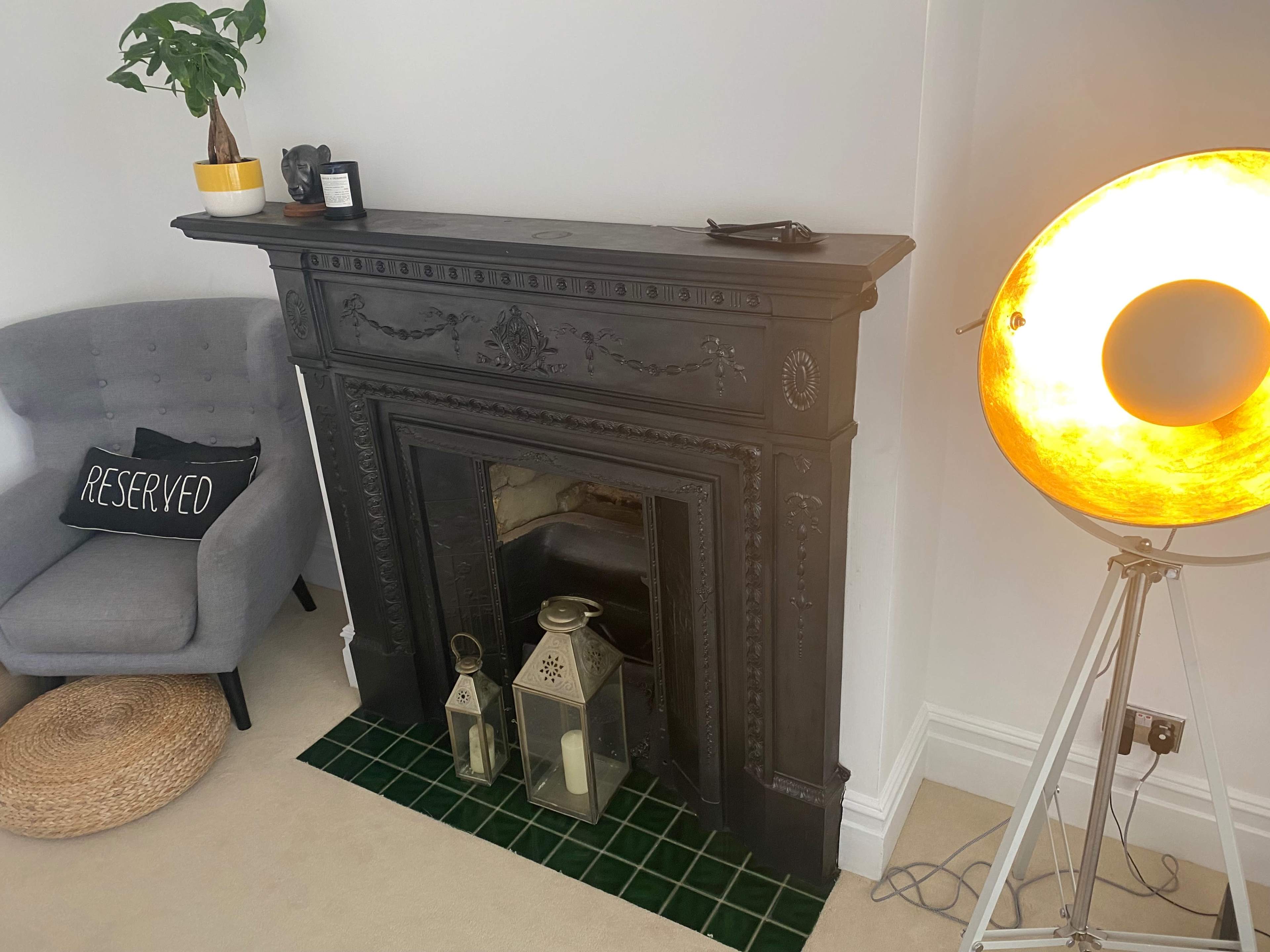 The image shows a black, ornate fireplace with two lanterns in front, a gray armchair with a "RESERVED" pillow, and a standing lamp with a glowing light.