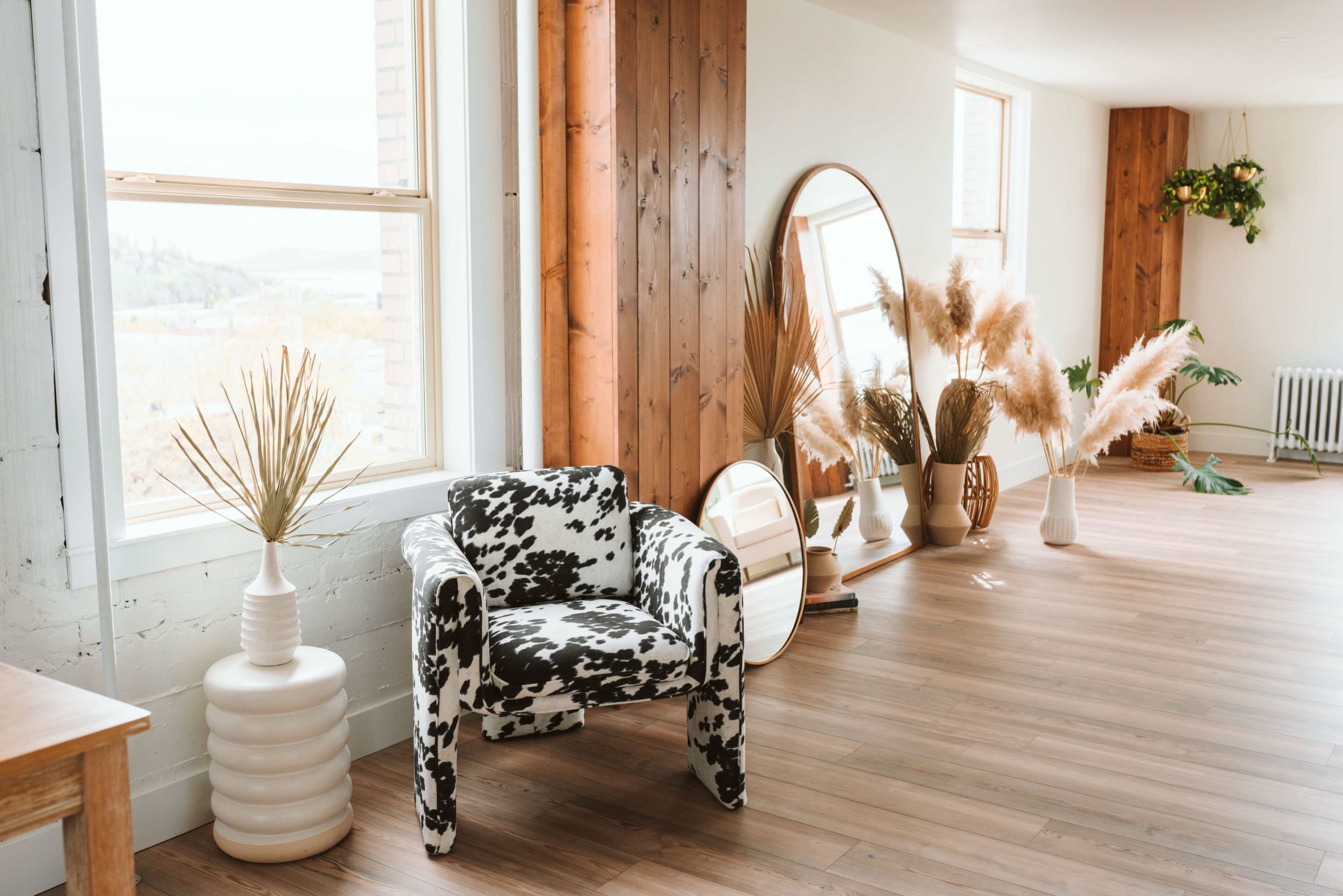 The image shows a spacious interior with a cow-print armchair, plants in decorative vases, and large windows allowing natural light to enter.