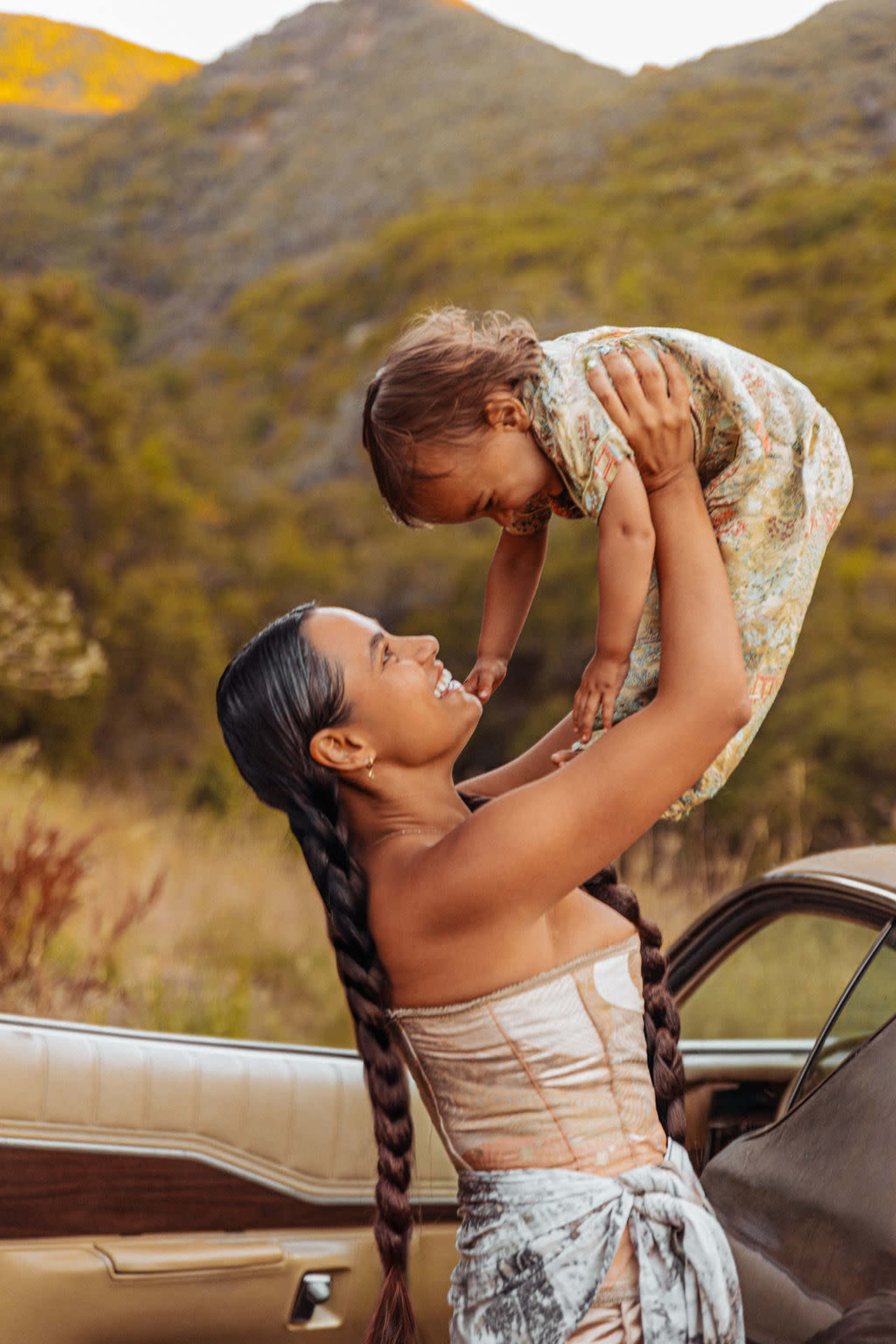 A woman holds a child above her head outside a car in a mountainous landscape.