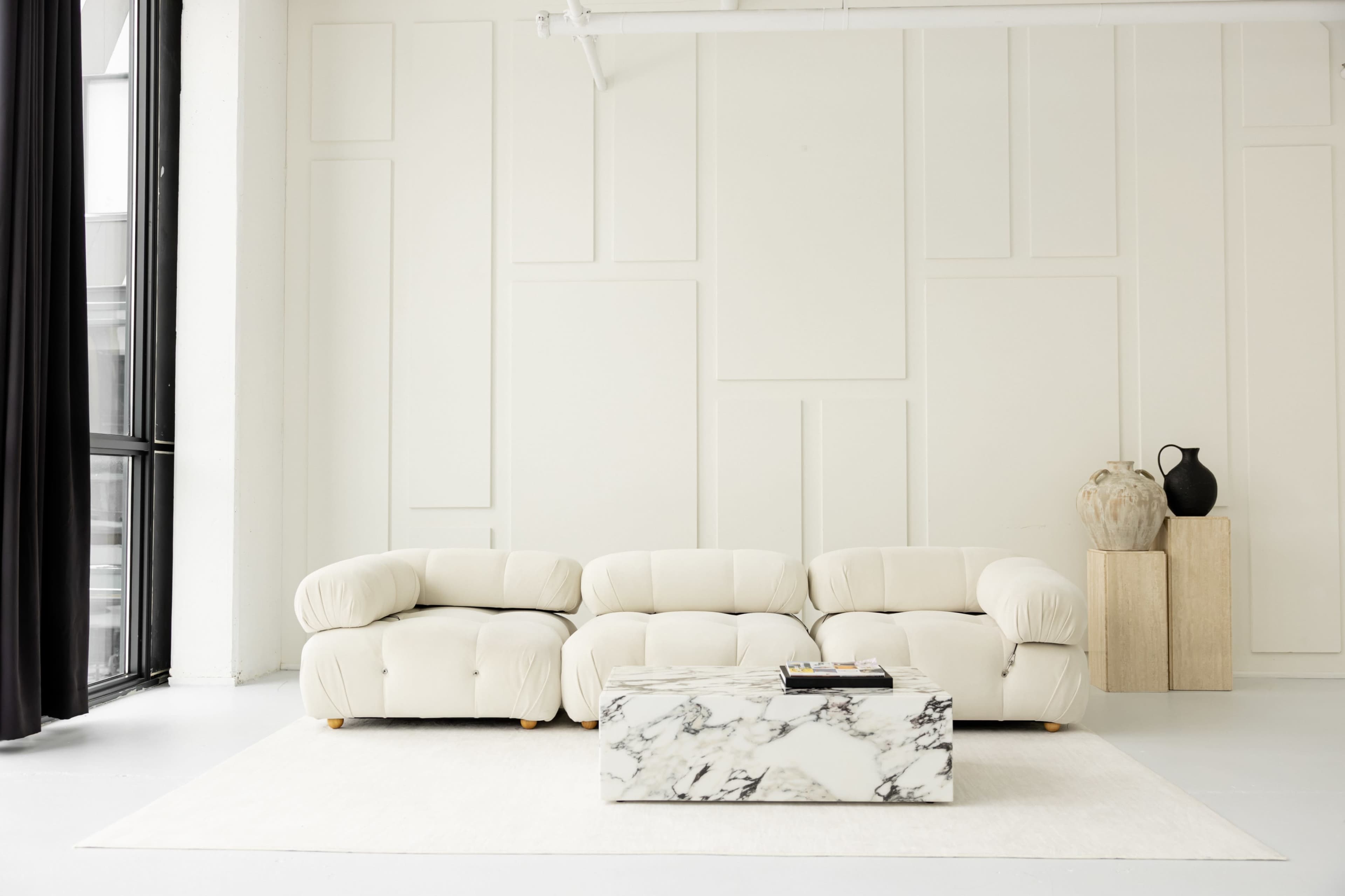 A light-colored, modern living room features a plush sectional sofa, a marble coffee table, and minimalistic decor against a neutral wall.