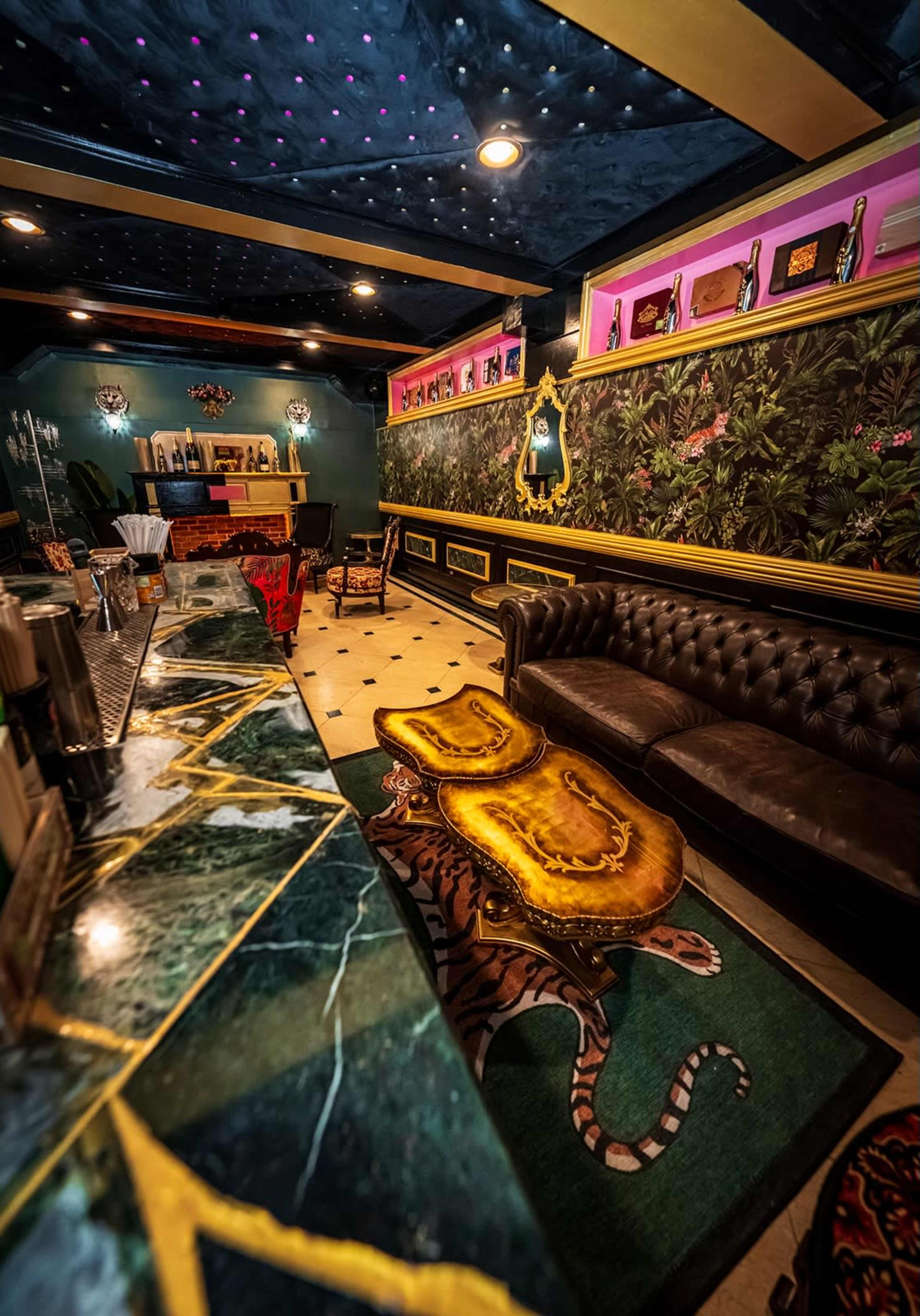 The image shows a stylish bar area featuring a dark leather sofa, a marble-topped counter, lush wallpaper, and unique decorative elements.