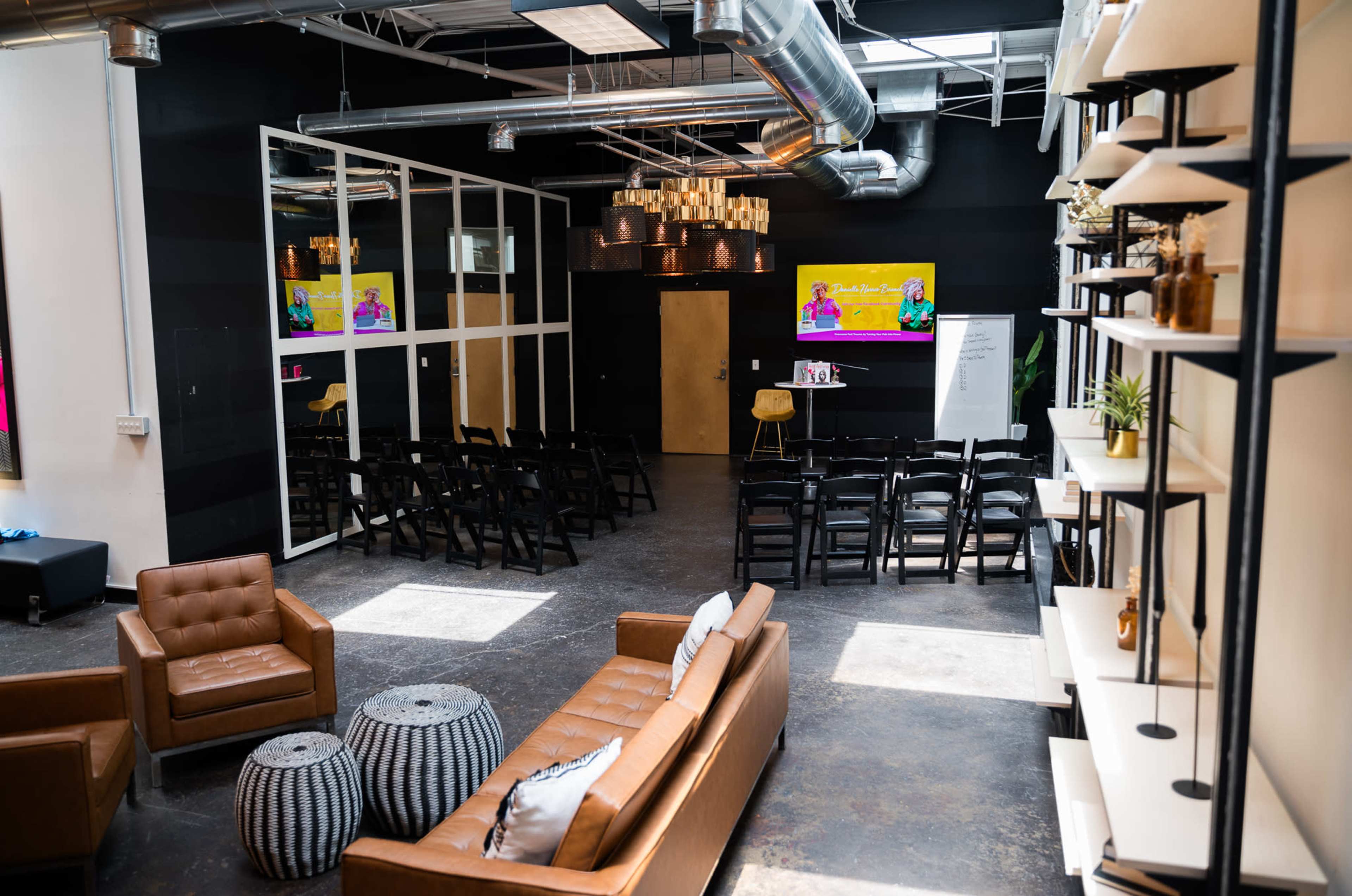 A modern, open event space featuring rows of chairs, a brown leather sofa, and two large screens displaying colorful presentations.