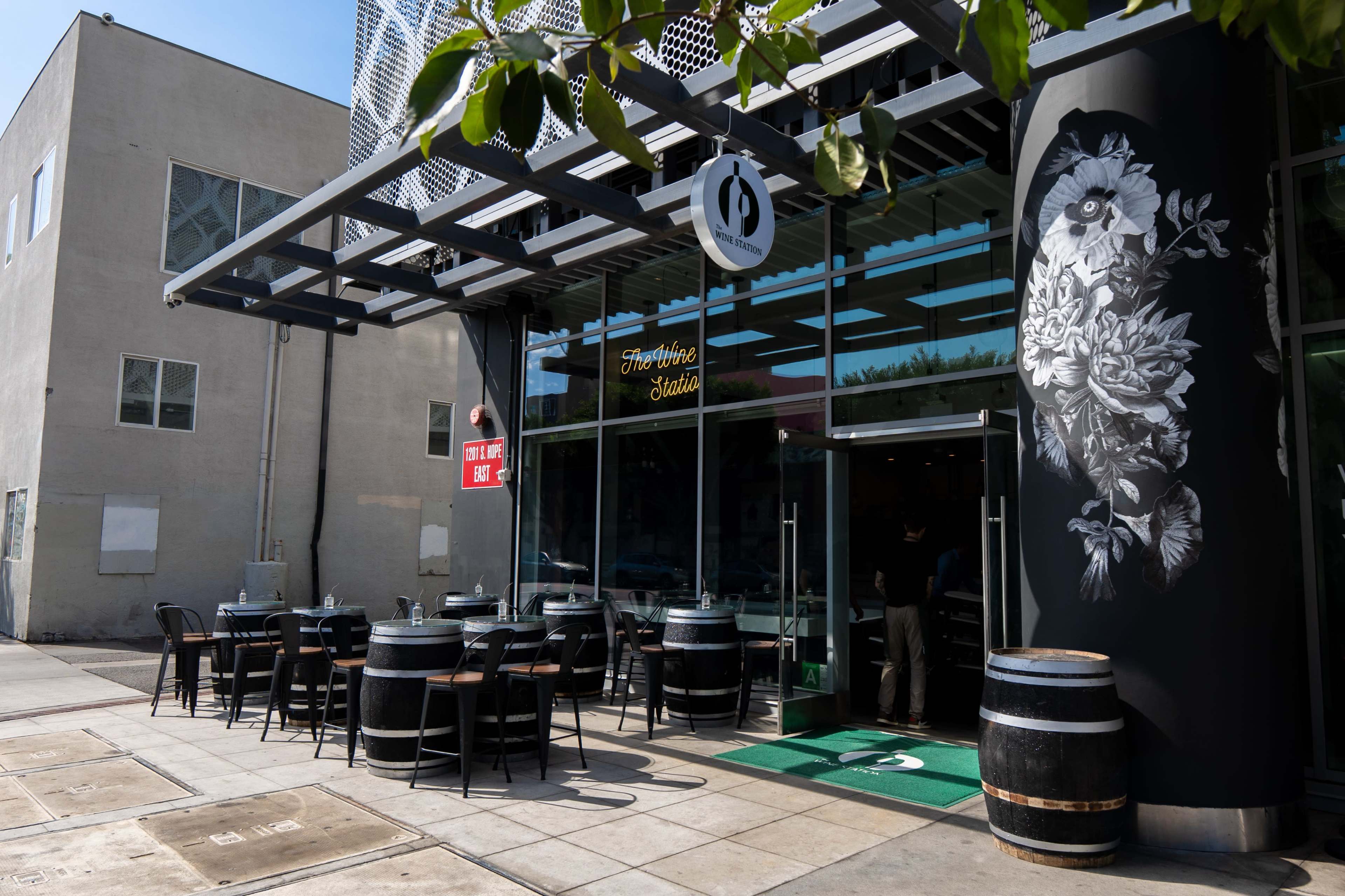 The image shows a modern café entrance featuring outdoor seating with wooden barrels and a large floral mural on the wall.