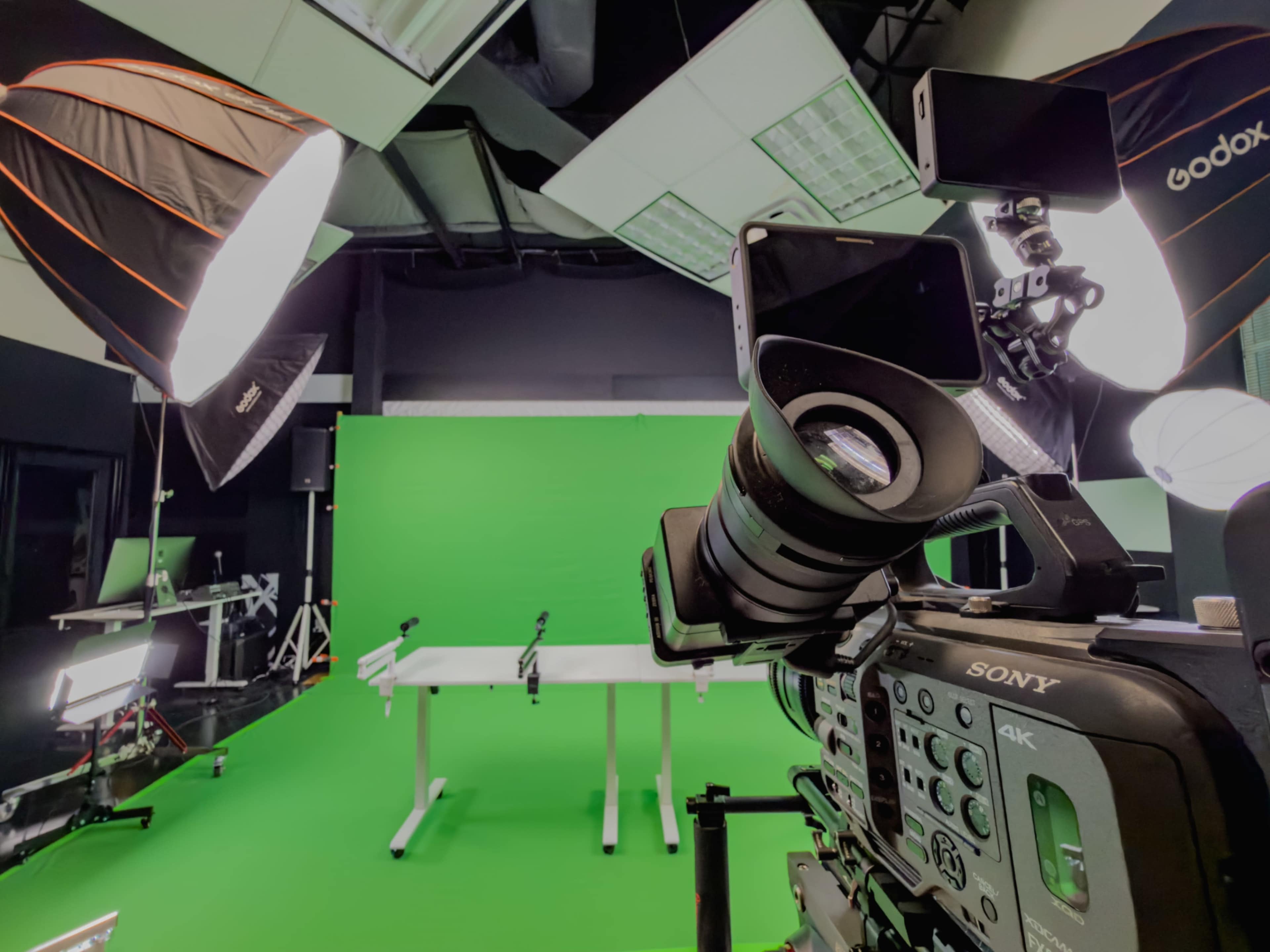 A camera is positioned in front of a green screen setup in a studio, with several lights and equipment arranged around a table.
