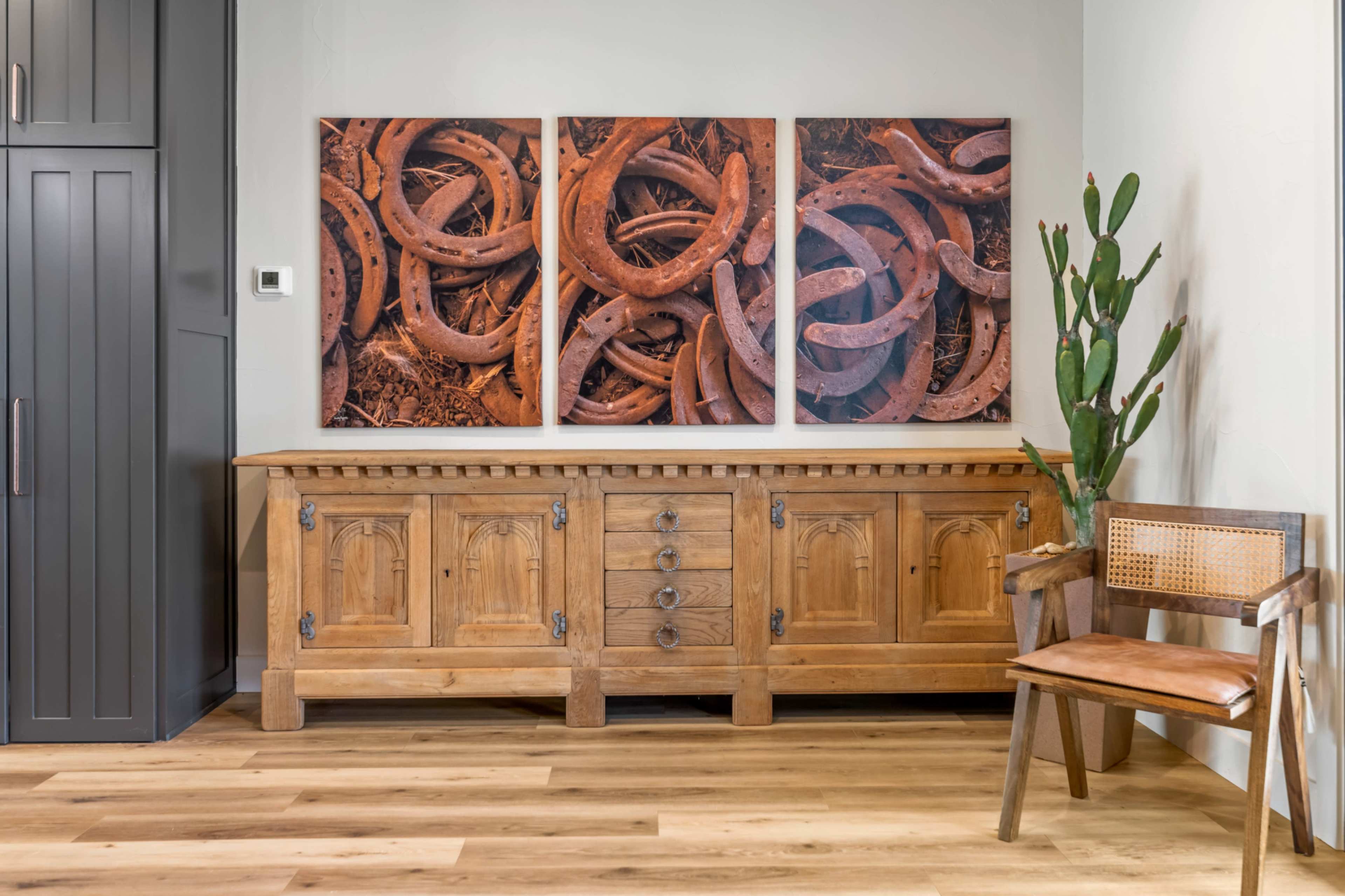 A wooden sideboard with intricate details is positioned against a wall featuring three large framed images of rusted metal rings, accompanied by a potted cactus and a wooden chair.