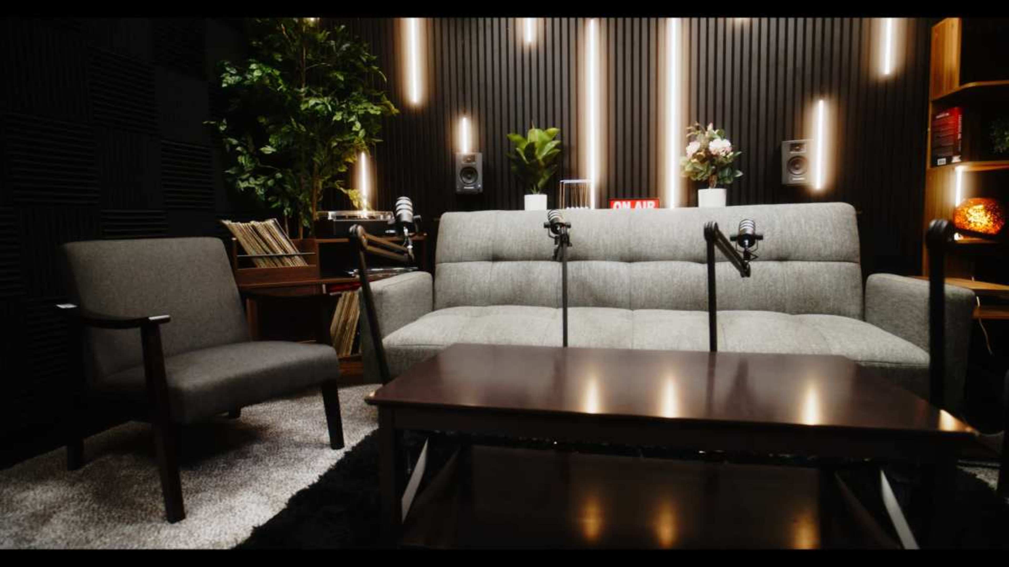 The scene shows a modern recording studio setup with a gray couch, two microphones on stands, a coffee table, and decorative plants along the walls.