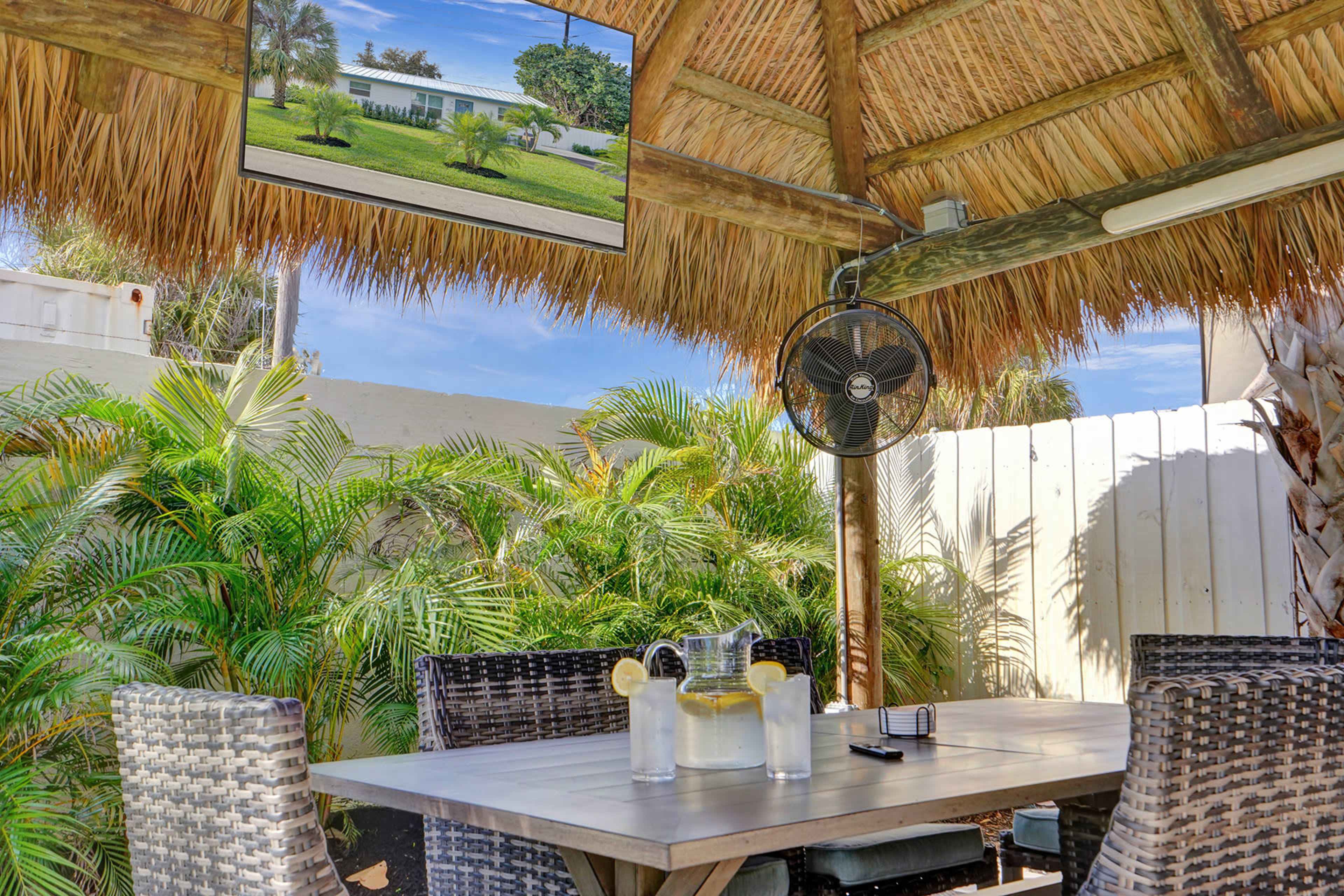 A thatched-roof patio features a dining table with chairs, a pitcher of lemonade, and a ceiling fan, surrounded by lush greenery.