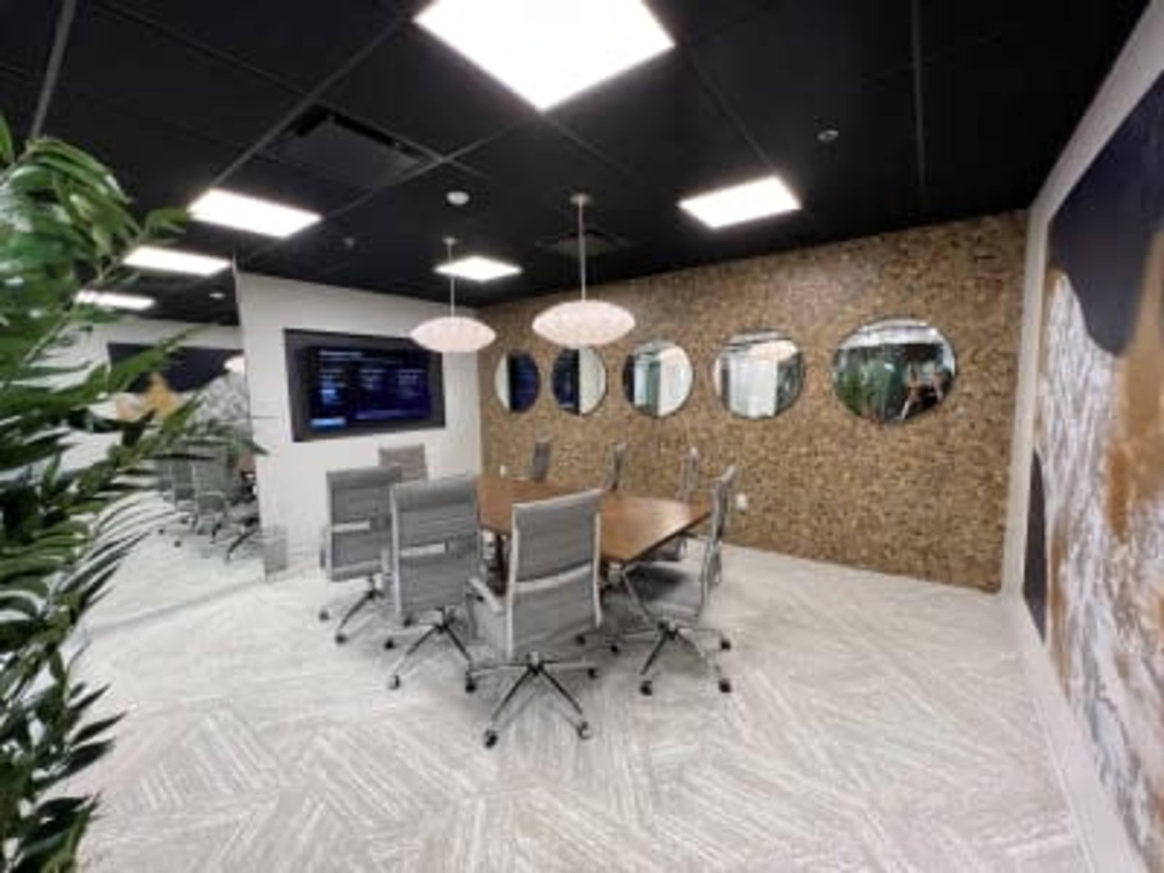 A modern conference room features a large table surrounded by gray chairs, with a wall of decorative mirrors and a display screen.
