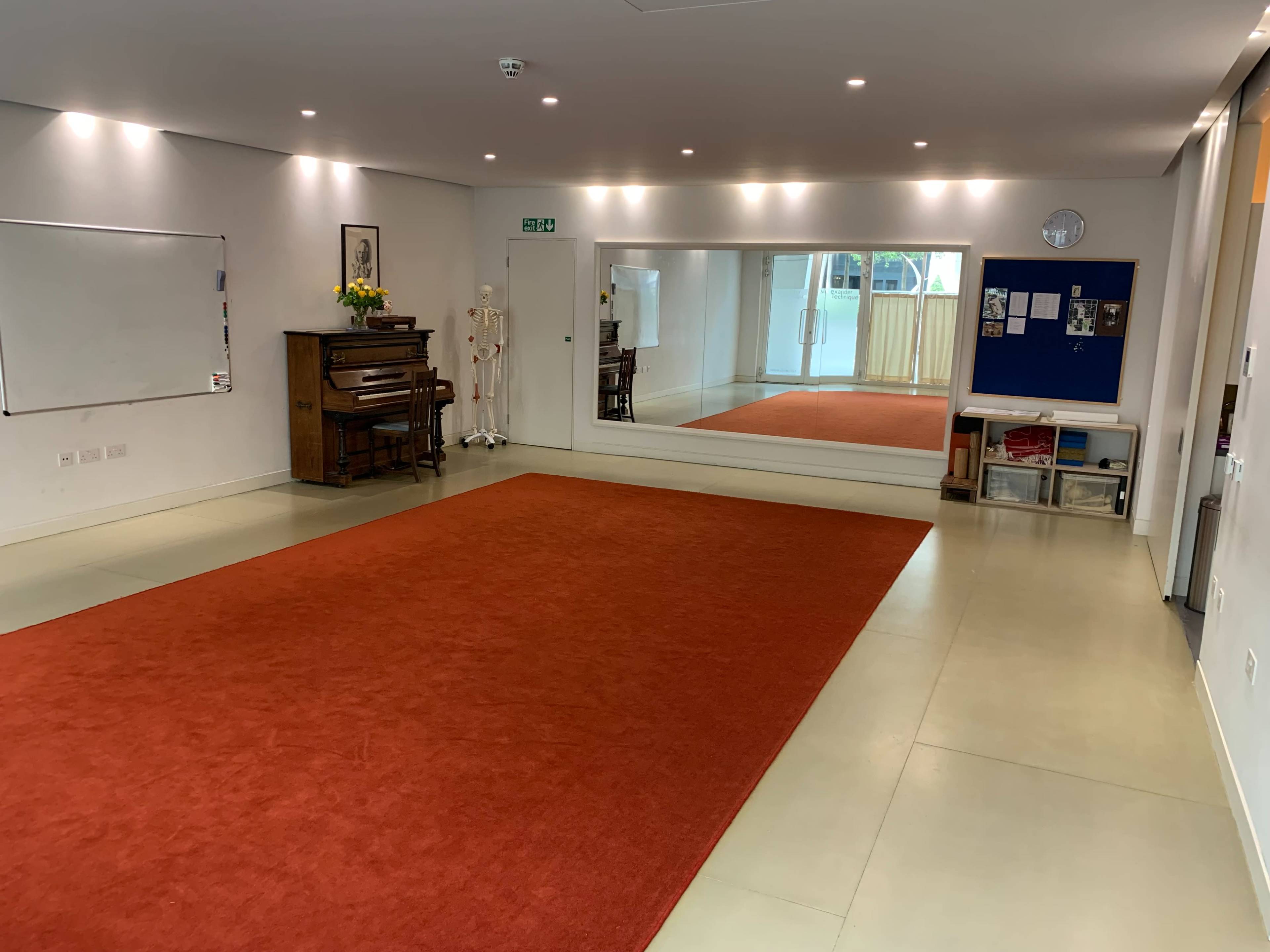 A spacious room with an orange carpet, a large mirror on one wall, a wooden piano, and a whiteboard.