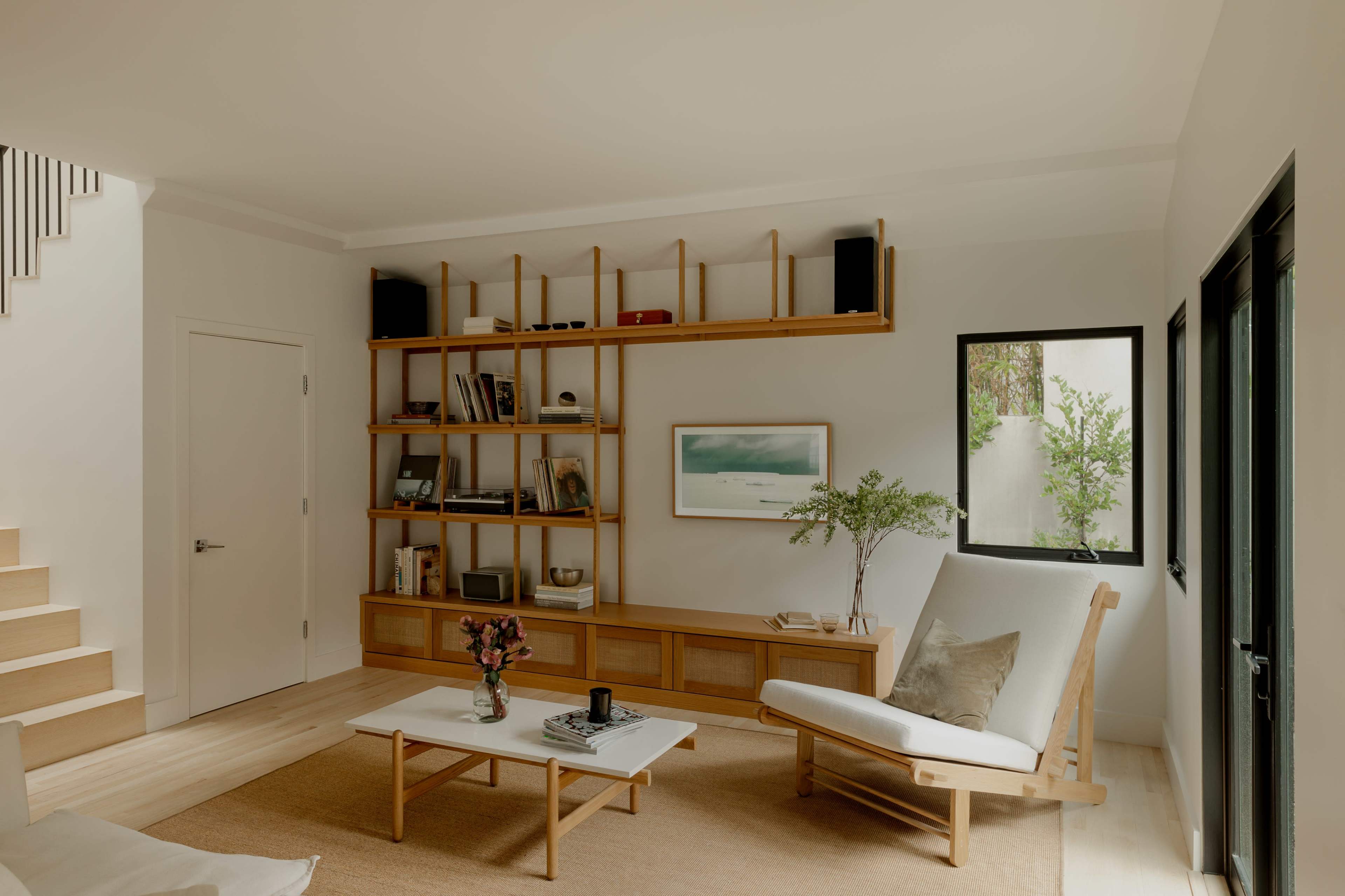 A minimalist living room with a wooden shelving unit, a white coffee table, a lounge chair, and a staircase in the background.