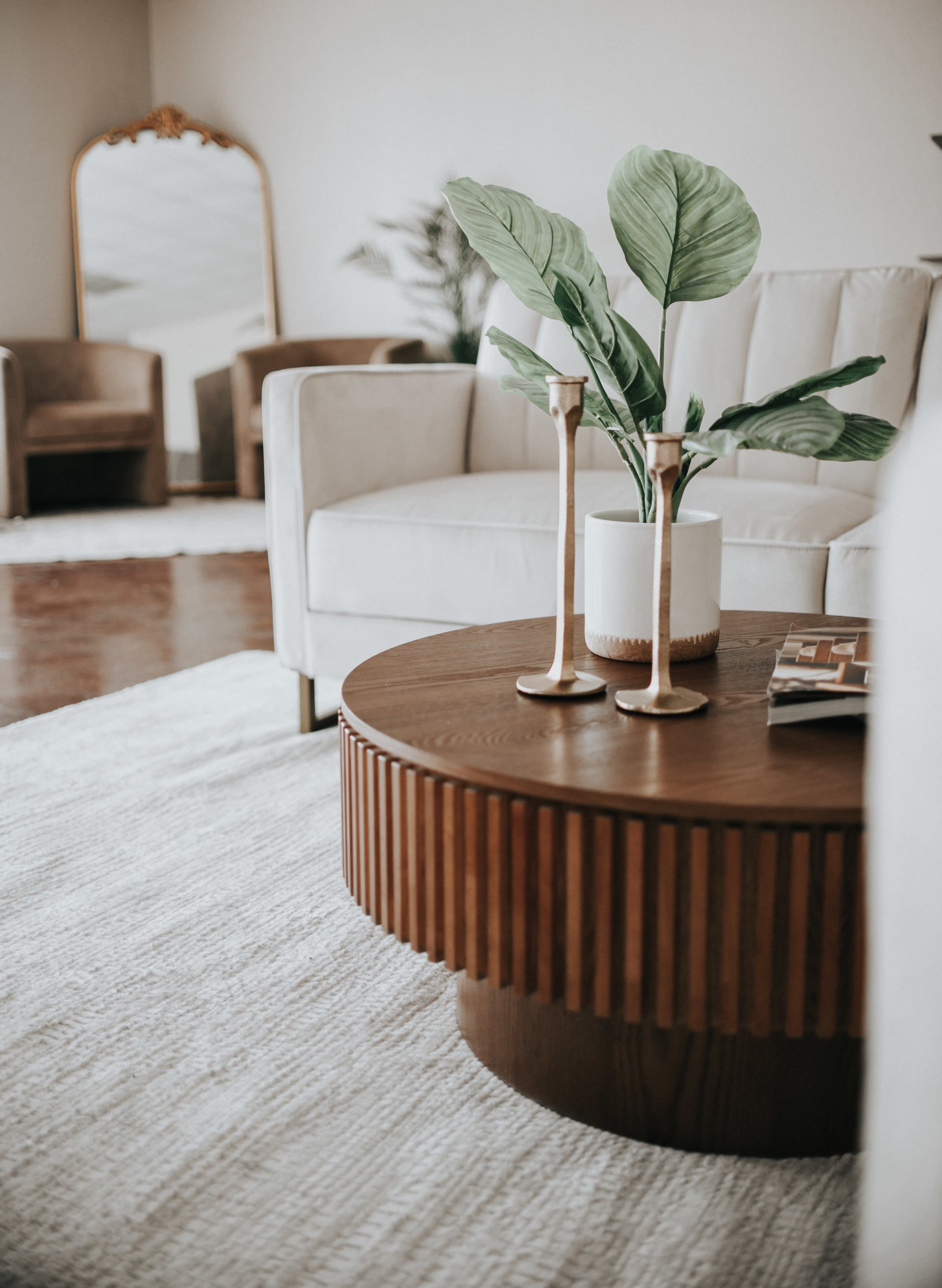 A wooden round coffee table with a textured base displays a potted plant and two candle holders in a modern living room setting.