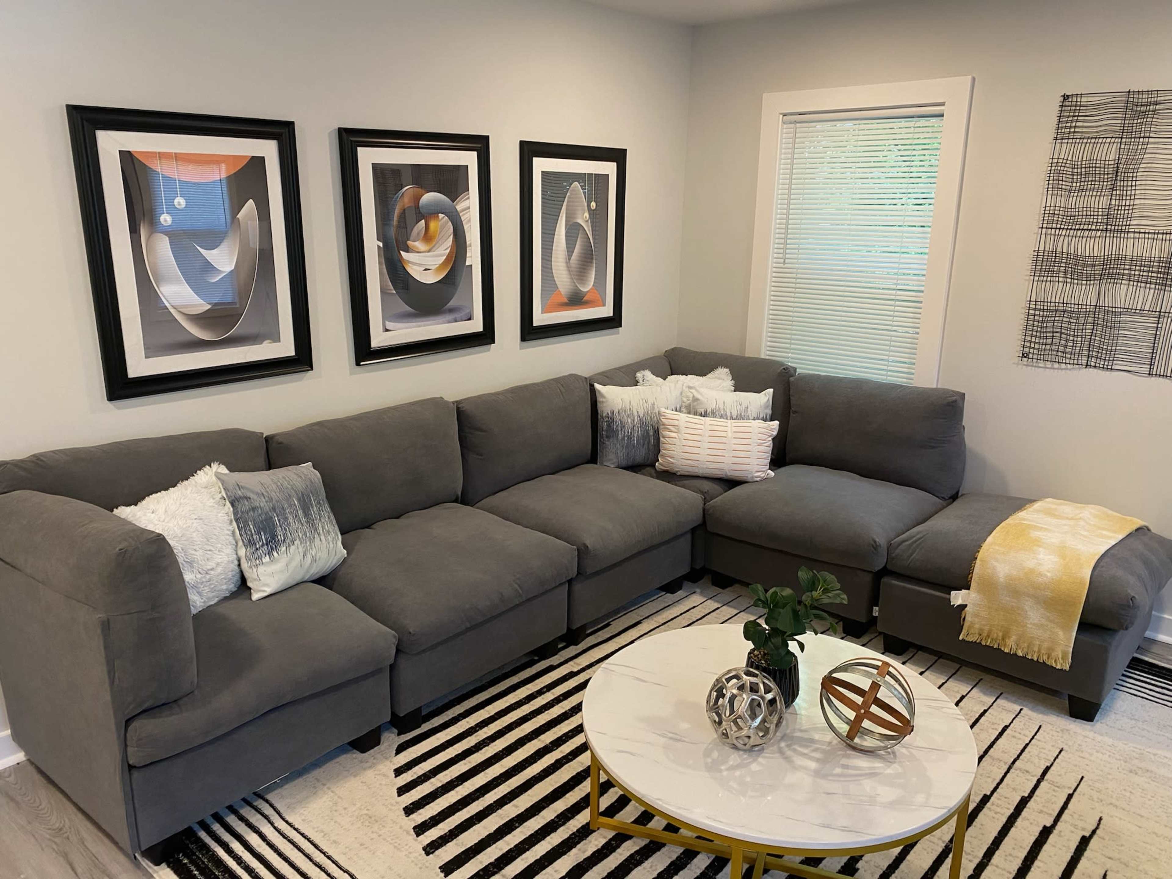 A gray sectional sofa is arranged against a wall with three framed abstract prints above it, accompanied by decorative pillows and a coffee table on a striped rug.