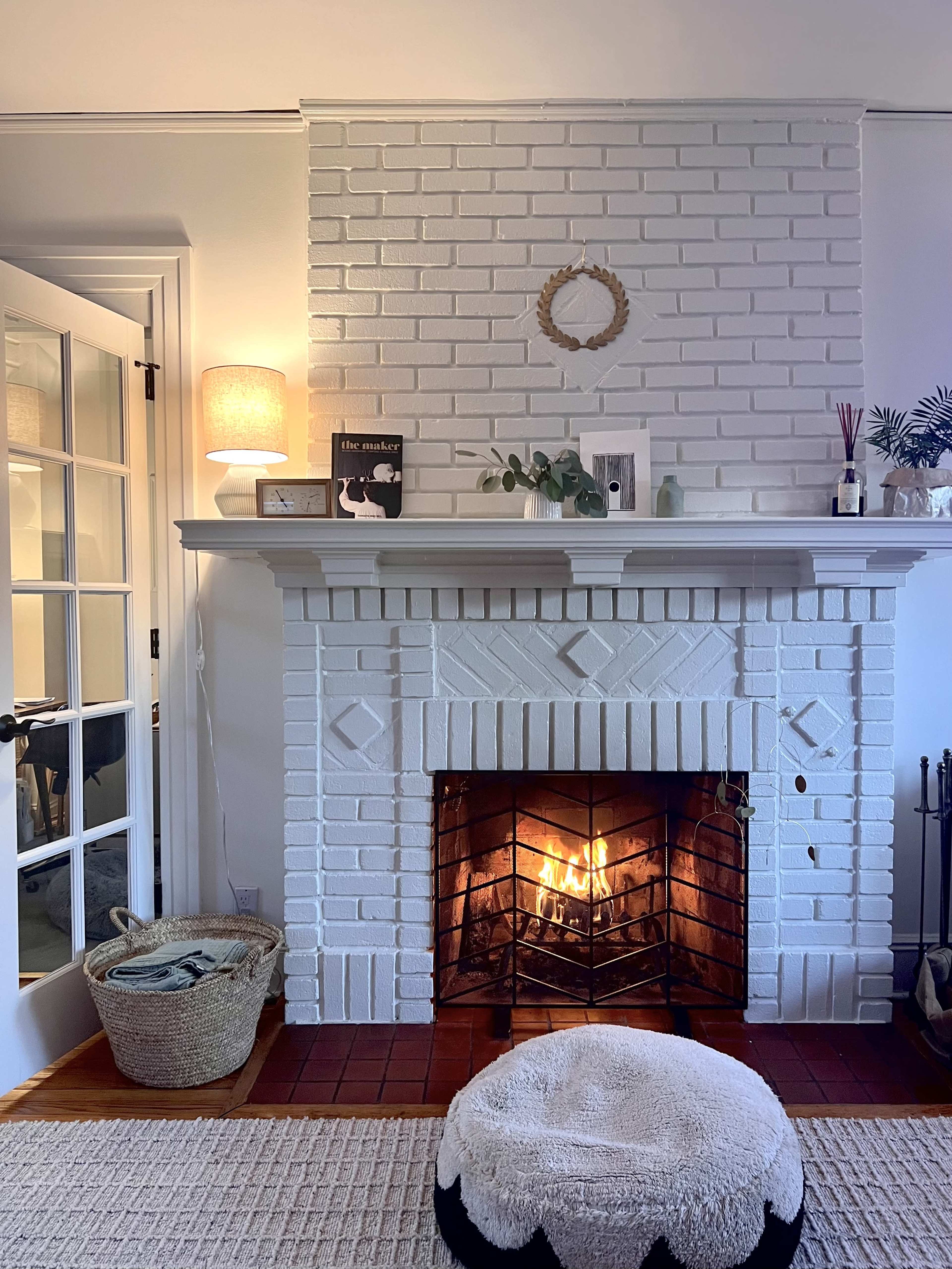 A white brick fireplace with a lit fire is surrounded by a decorative mantel, a large woven basket, and a round pouf, all in a cozy living room setting.