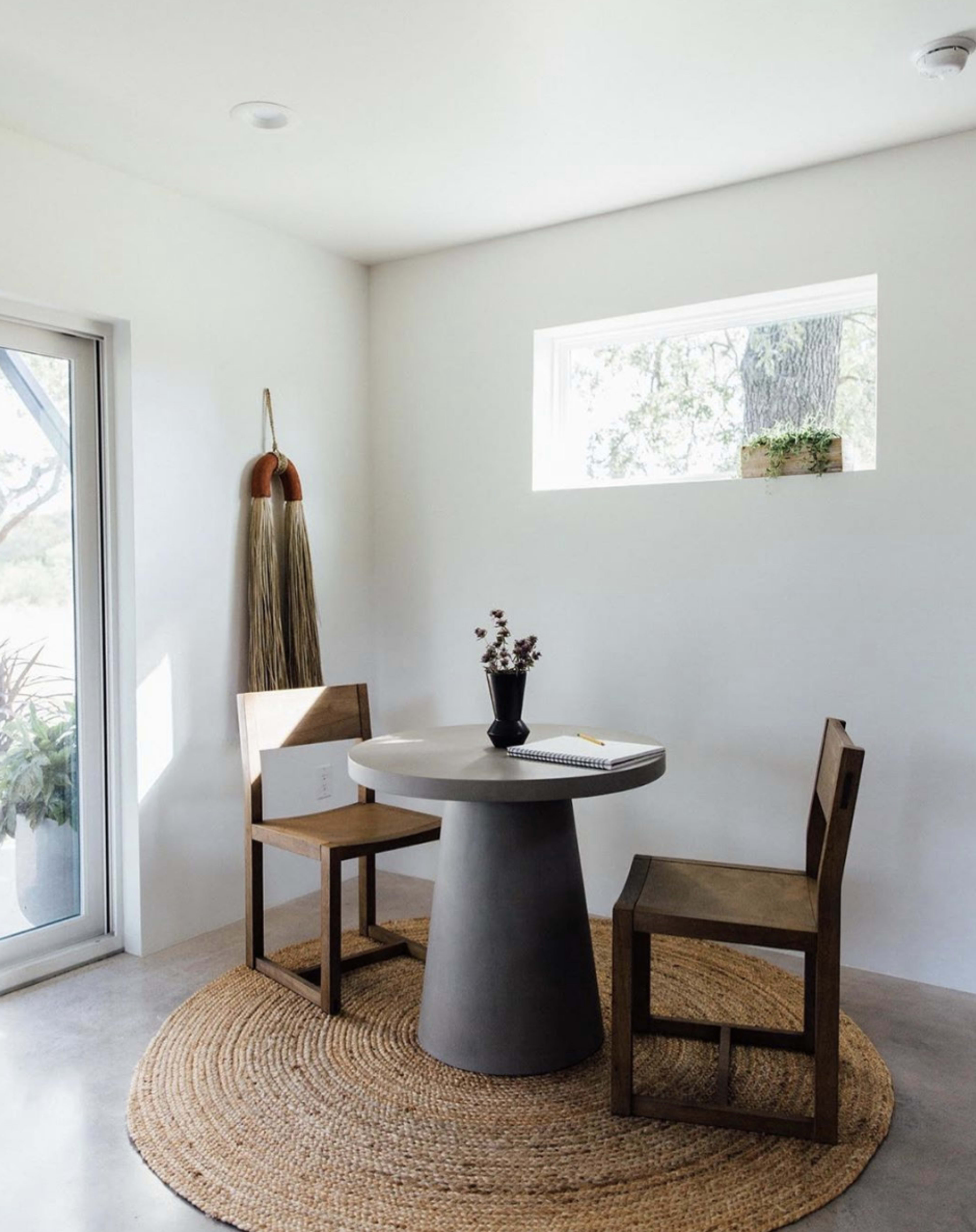 A round table with two wooden chairs sits on a woven rug beneath a small window in a minimalist, well-lit room.
